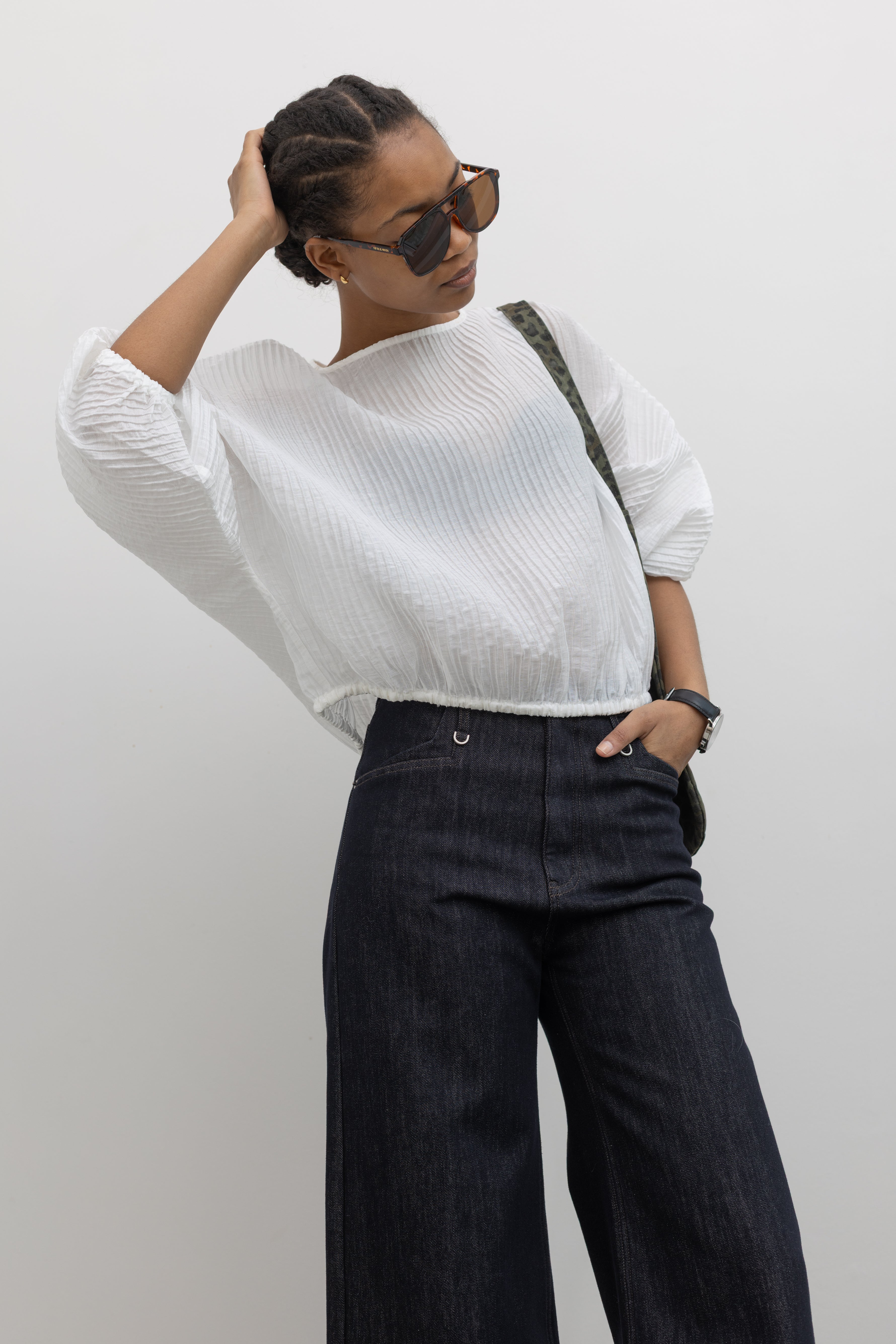 Front view of a woman standing wearing a lightweight flowy blouson tencel top with a relaxed drape and subtle pleated texture and a side tie at the hem paired with dark wash denim jeans at Alas Eius.