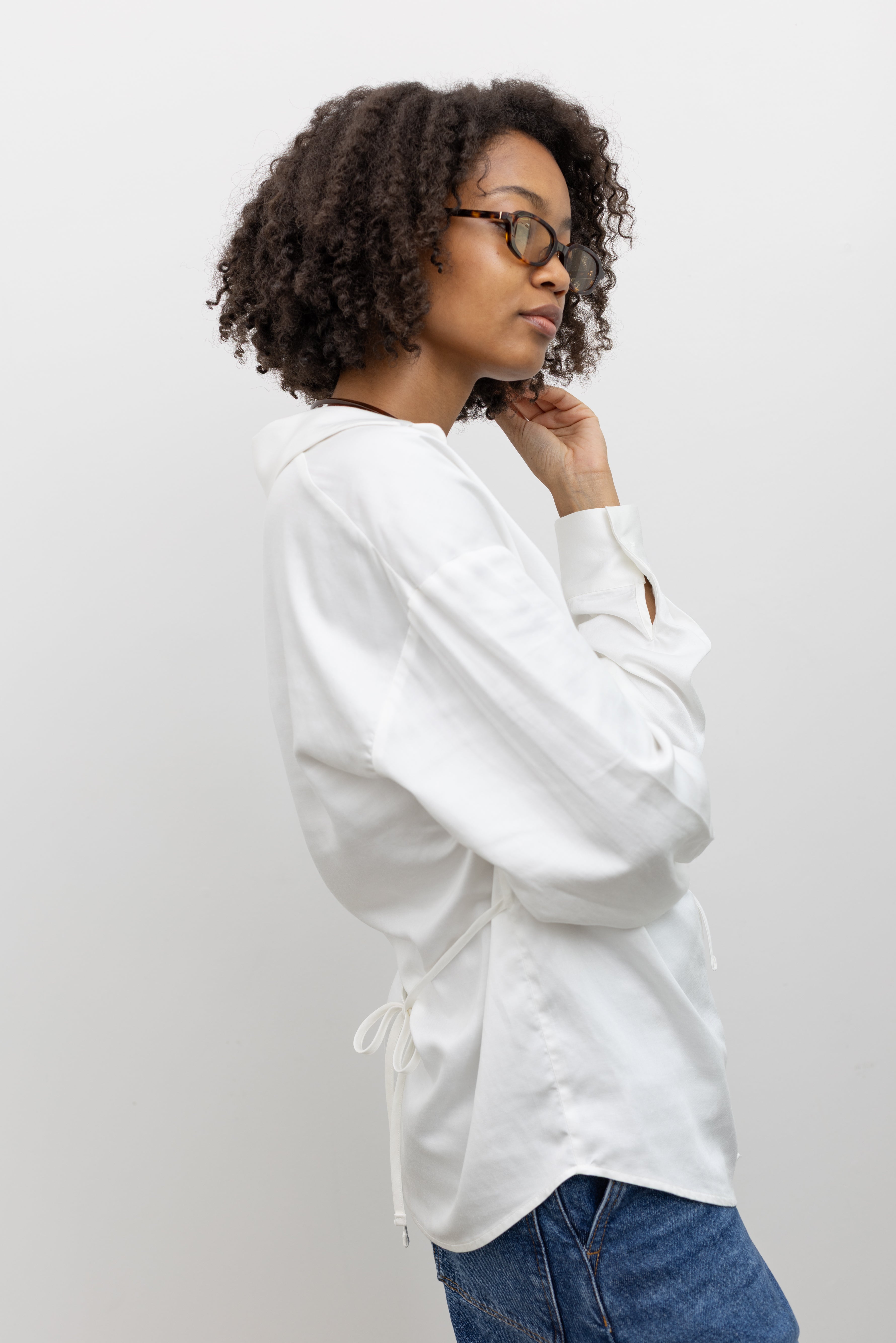 Woman wearing modern minimalist summer fashion in a Collective lightweight white cotton gathered blouse with long sleeves, a relaxed fit, and front tie detail, and deep v neckline paired with blue denim jeans at Alas Eius. 