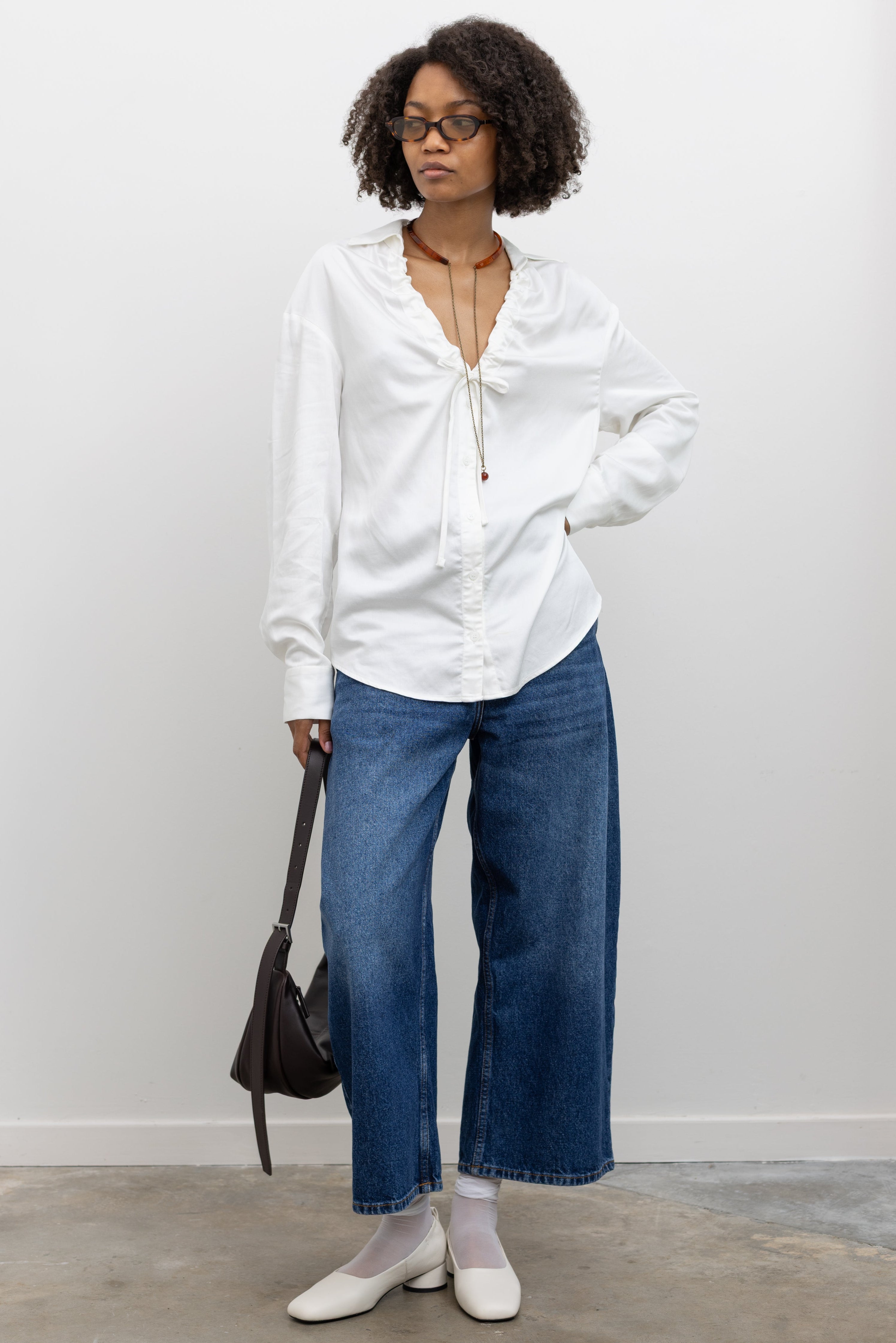 Woman wearing chic summer fashion in a Collective lightweight white cotton gathered blouse with long sleeves, a relaxed fit, and front tie detail, and deep v neckline paired with cropped ankle length blue denim jeans and white pumps at Alas Eius. 