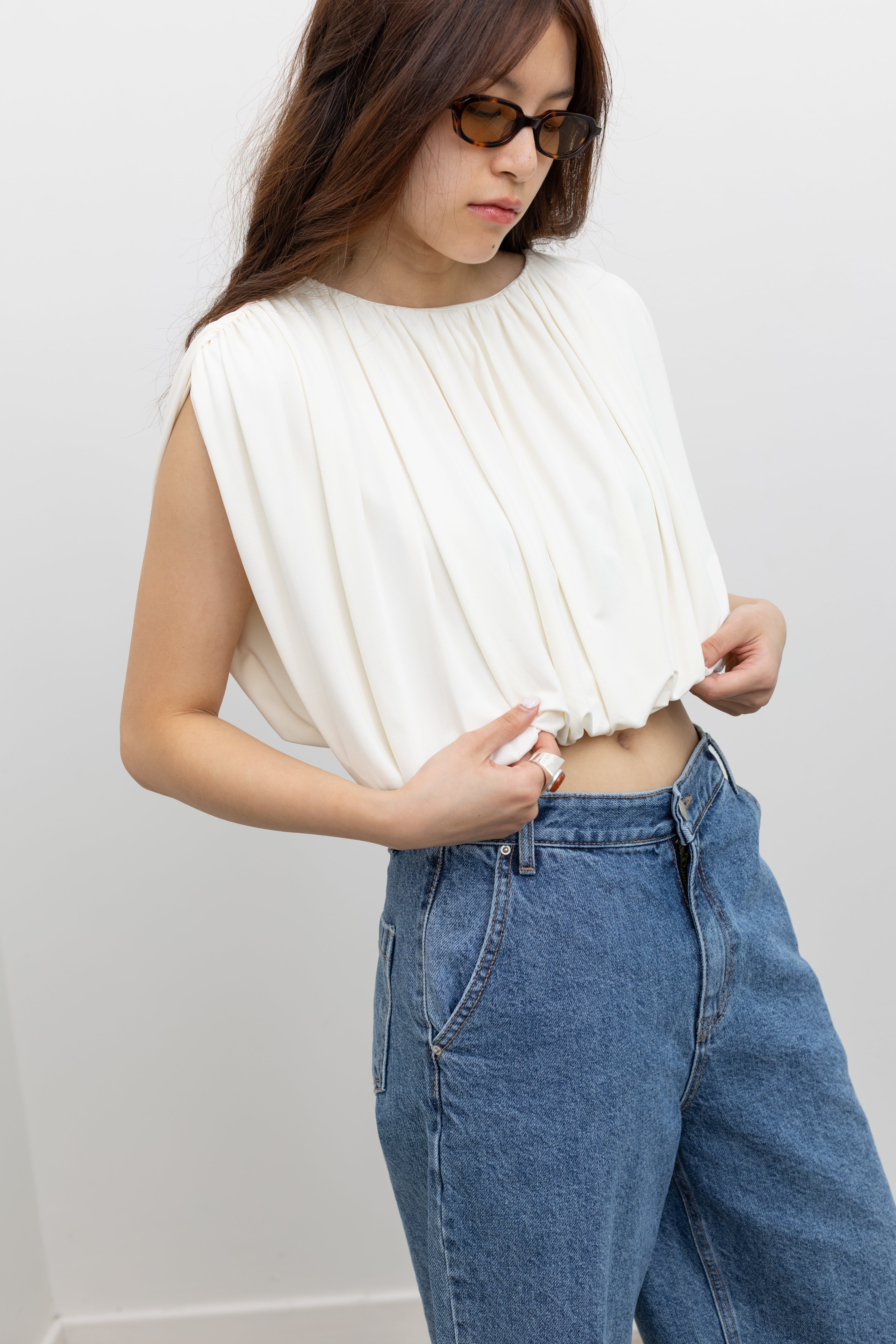 Model wearing chic summer fashion in a Collective white pleated balloon top with all-over gathers, a voluminous puff cropped hem, and a fully lined relaxed silhouette paired with stone washed bermuda shorts at Alas Eius. 