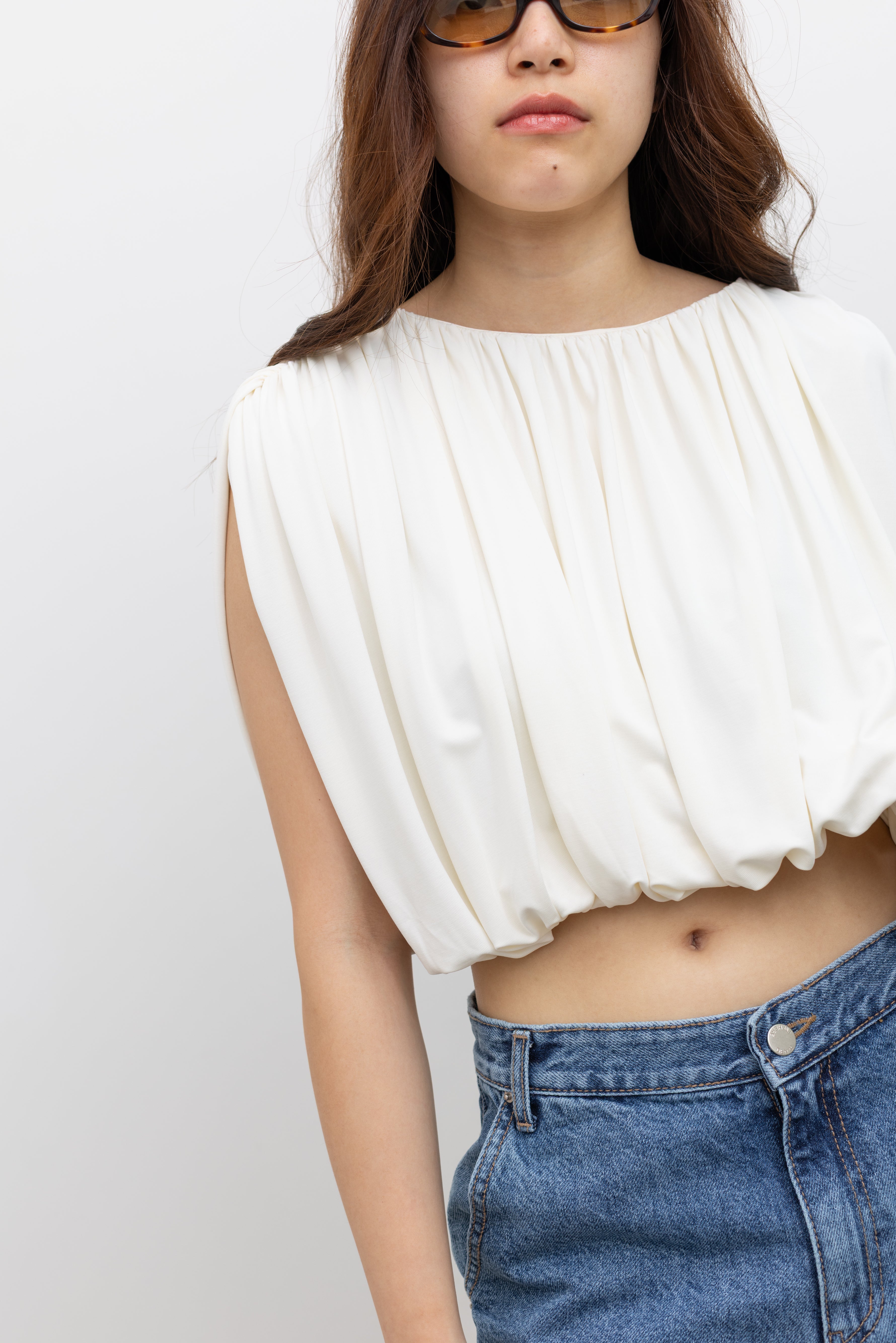 Model wearing elevated summer fashion in a Collective white pleated balloon top with all-over gathering, a voluminous puff hem, and a fully lined relaxed silhouette paired with stone washed bermuda shorts at Alas Eius. 