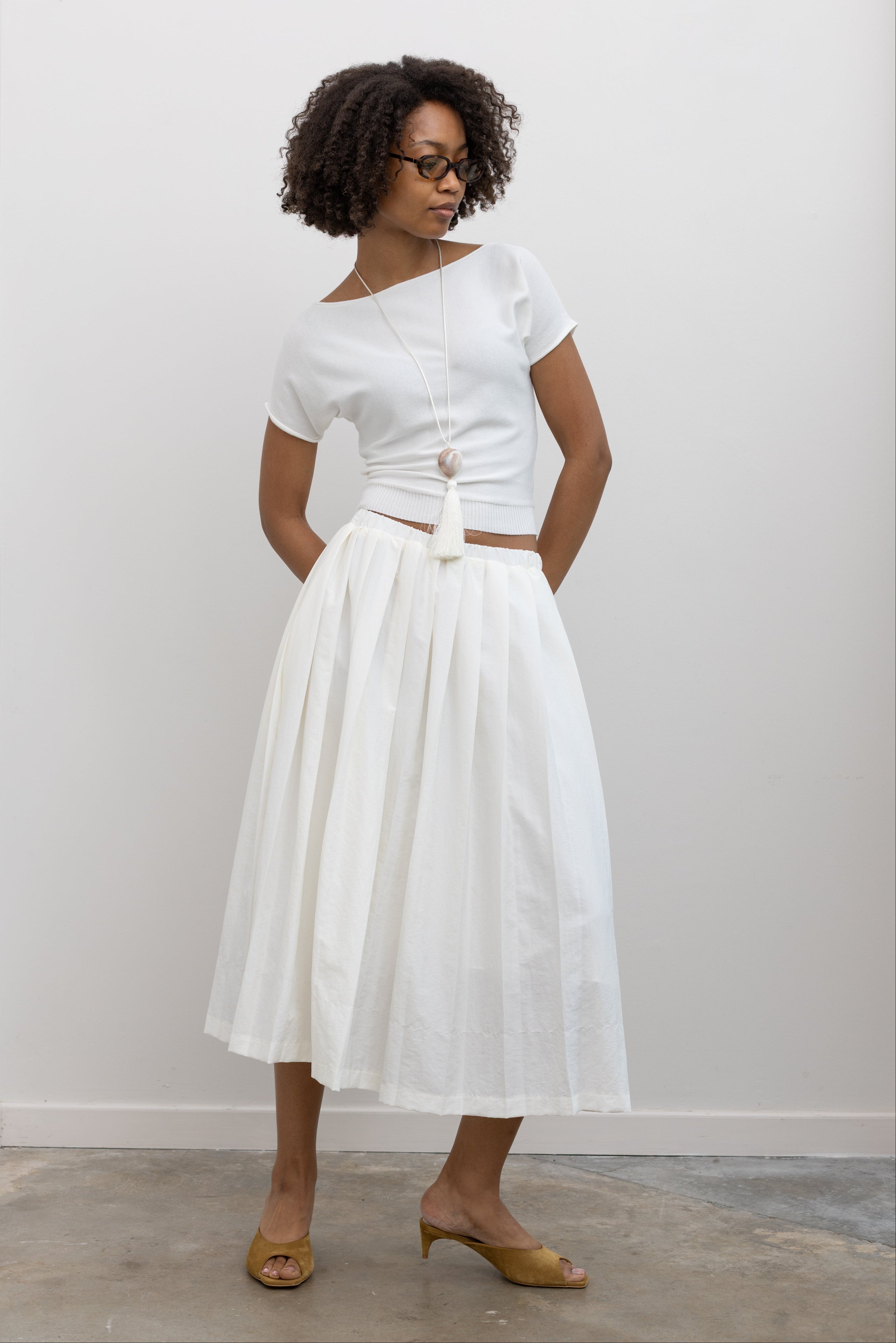 Woman wearing chic summer fashion in a Collective brand ivory knit boat neck cinched tee paired with an Alas Eius label white nylon pleated midi skirt at Alas Eius. 