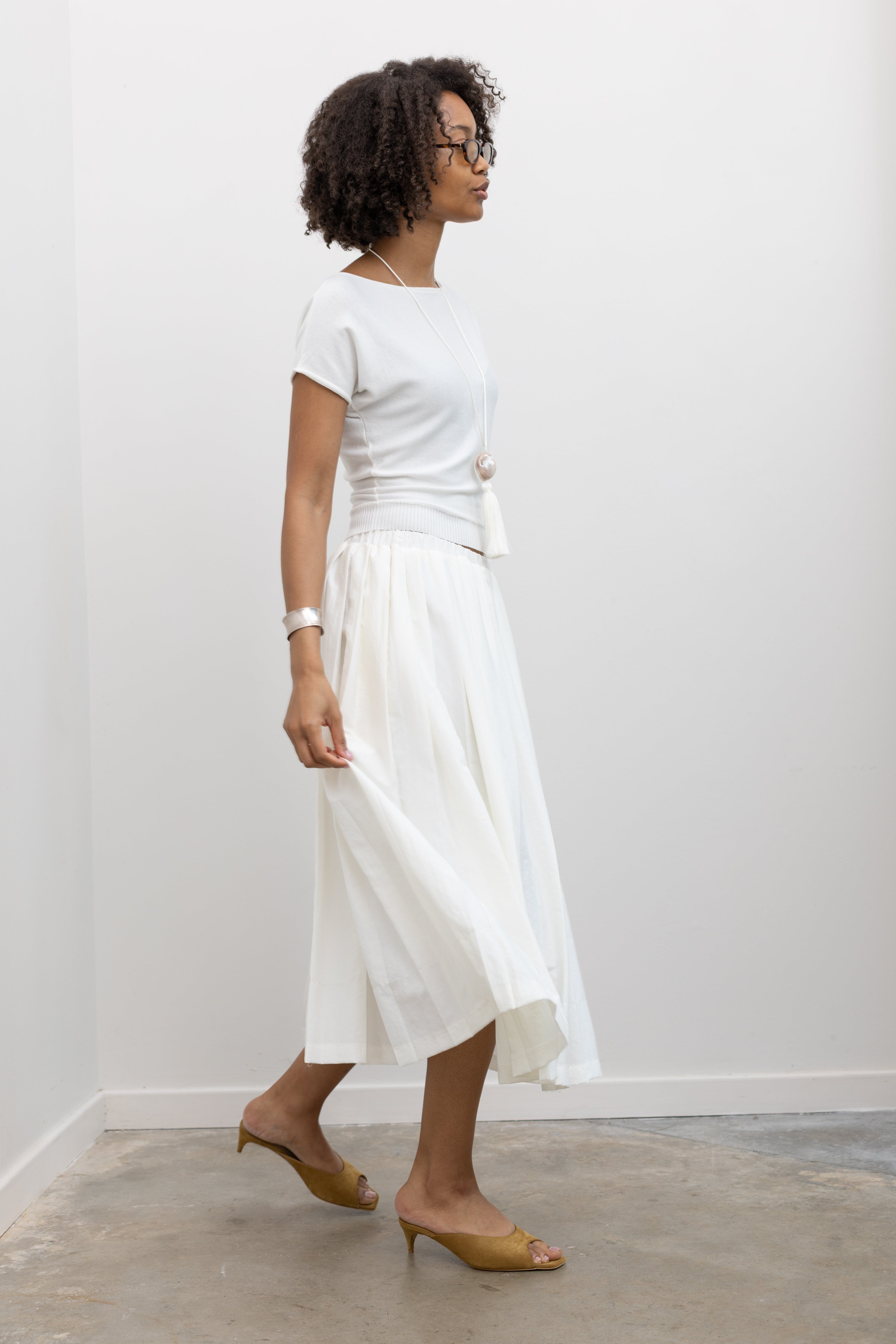 Woman wearing sophisticated summer fashion in a Collective brand ivory knit boat neck cinched tee paired with an Alas Eius label white nylon pleated midi skirt at Alas Eius. 