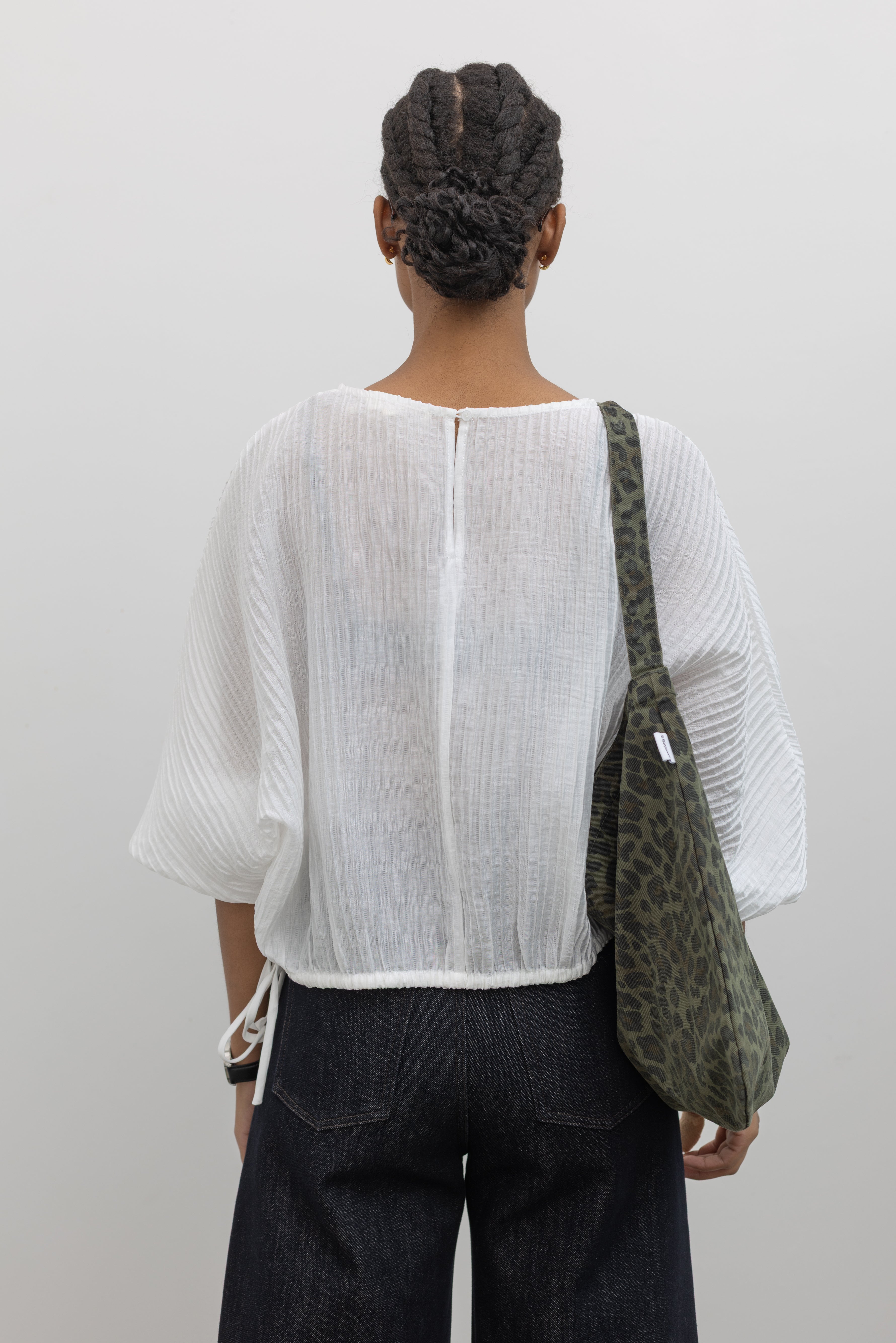 Product back view of a woman standing wearing a lightweight flowy blouson tencel top with a relaxed drape and subtle pleated texture and a side tie at the hem paired with dark wash denim jeans at Alas Eius.