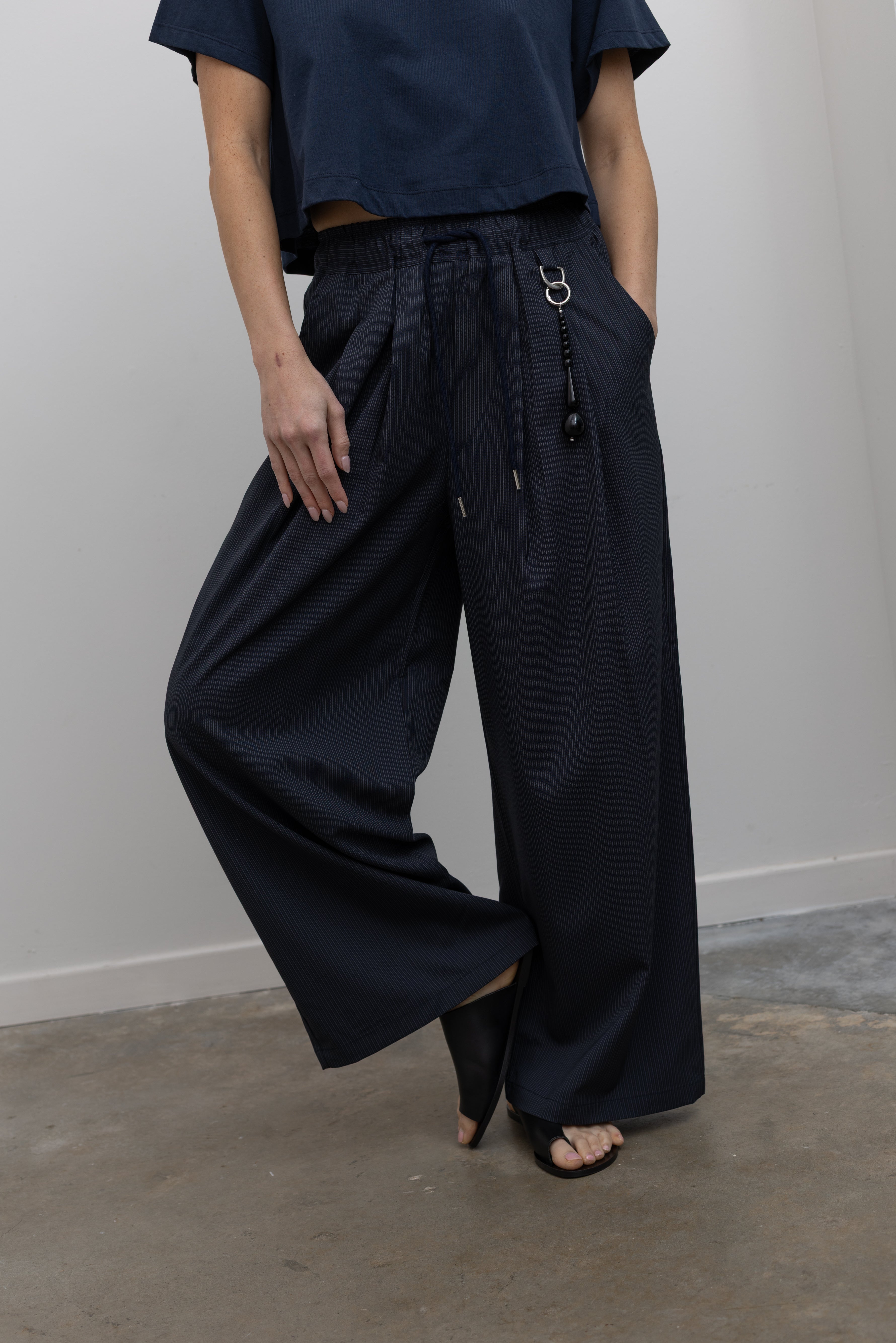 Detailed product view of Woman standing wearing Spring fashion in a pair of Collective Navy Pinstripe Wide Leg Drawstring Pants and navy blue tee with black beaded accessory at belt loop at Alas Eius