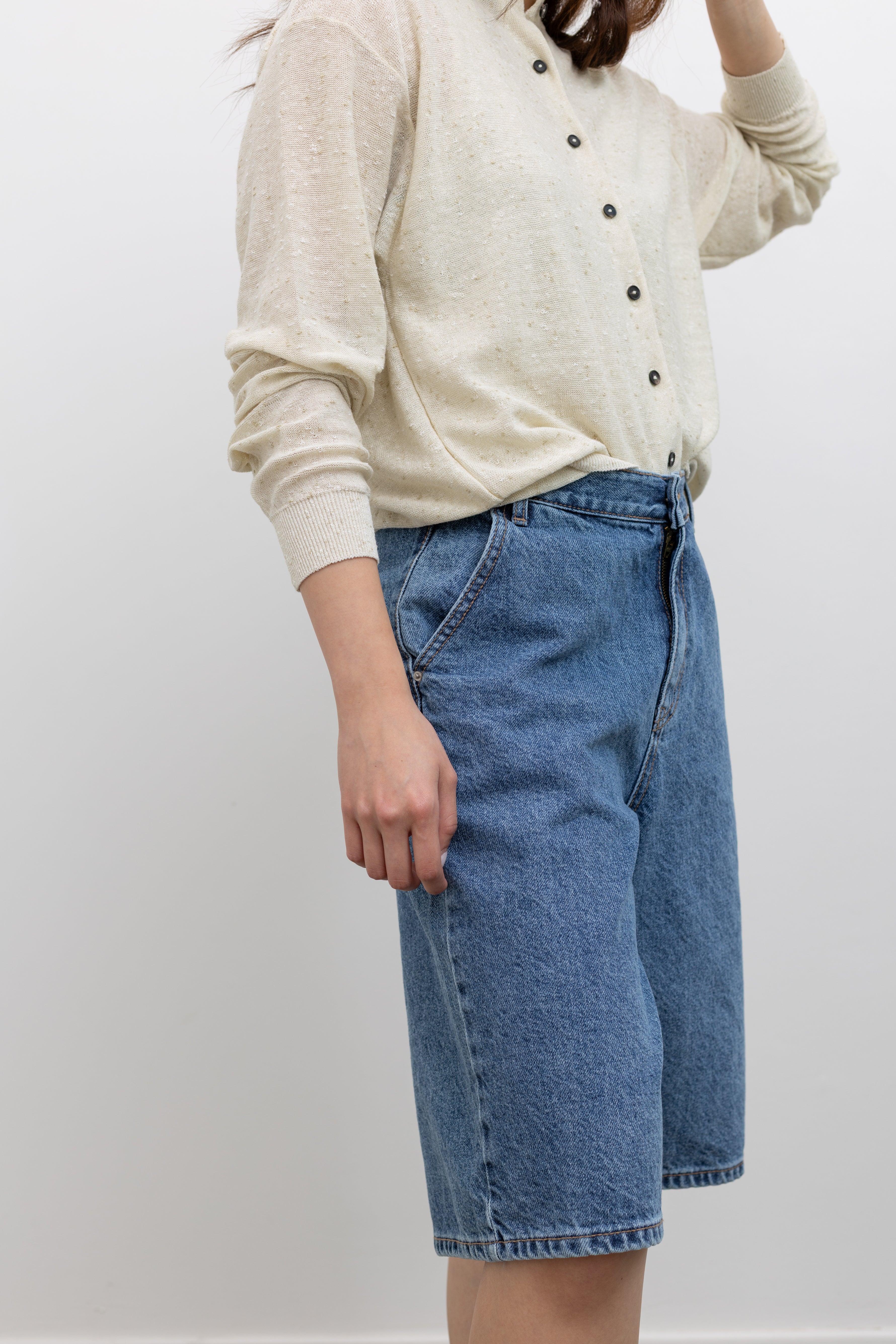 Model wearing chic summer fashion in the Collective stone-washed light blue denim bermuda shorts with a mid-to-high rise, wide-leg silhouette, and knee-length design paired with a Cordera cotton linen blend cardigan and brown wooden beaded bucket hat at Alas Eius.
