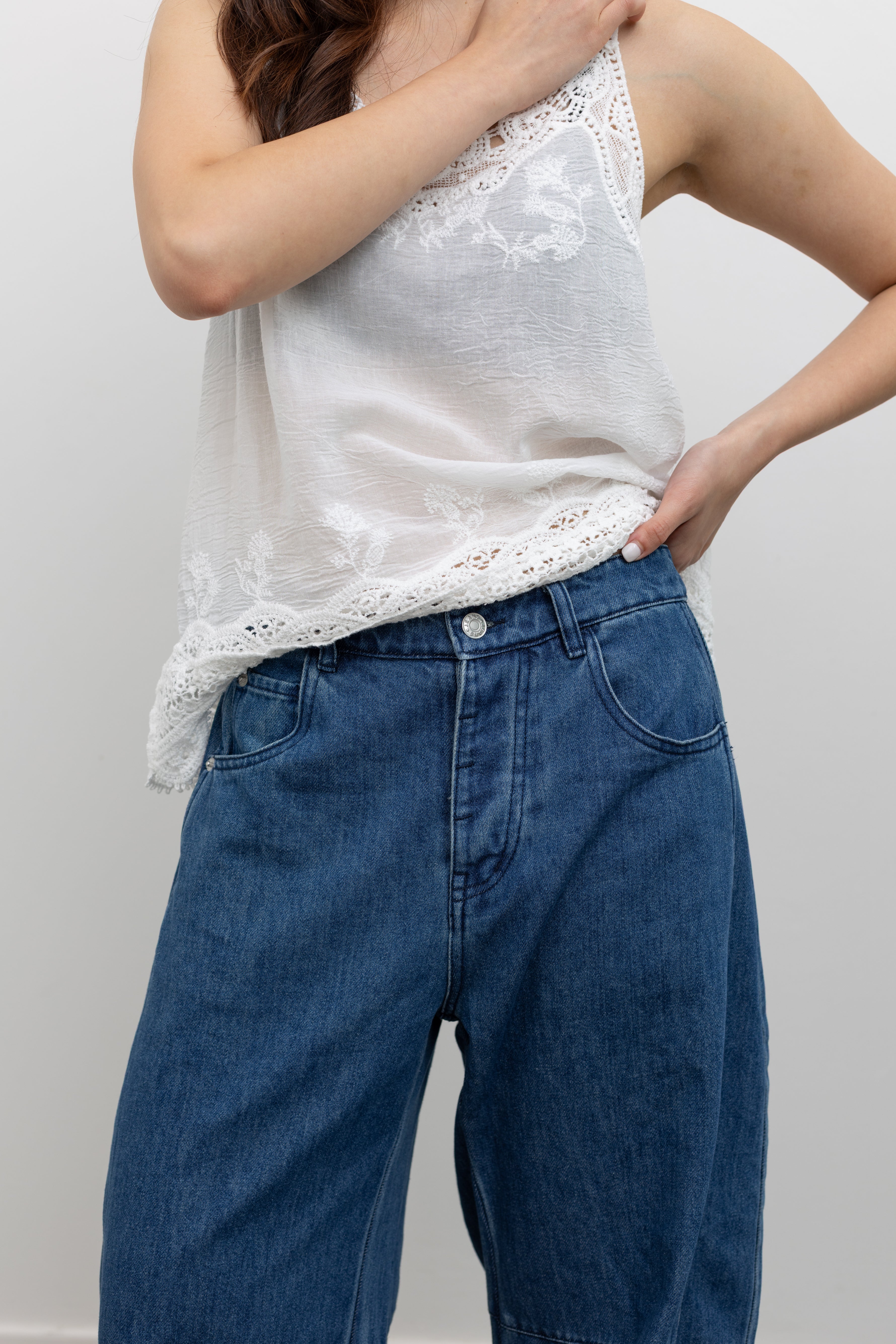 Model wearing modern baggy jeans in the Collective blue cotton baggy denim pants with curved balloon silhouette, mid-rise waist, and full-length hem paired with a crochet lace camisole tank top and Veja blue suede sneakers at Alas Eius. 