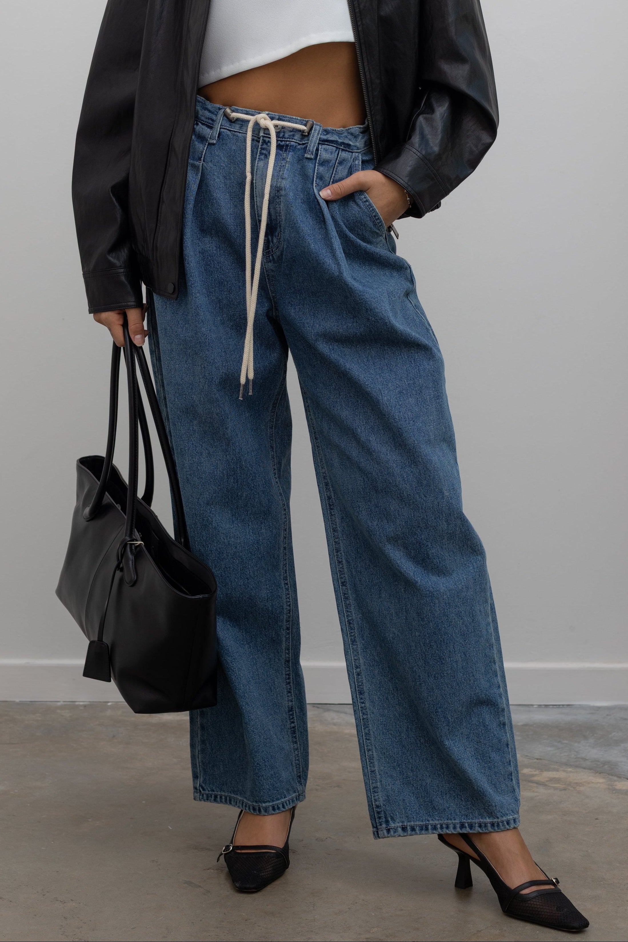 Woman wearing pleated front jeans featuring a drawstring waist, a mid-rise silhouette, and a straight leg cut paired with slingback heels and a leather jacket at Alas Eius. 