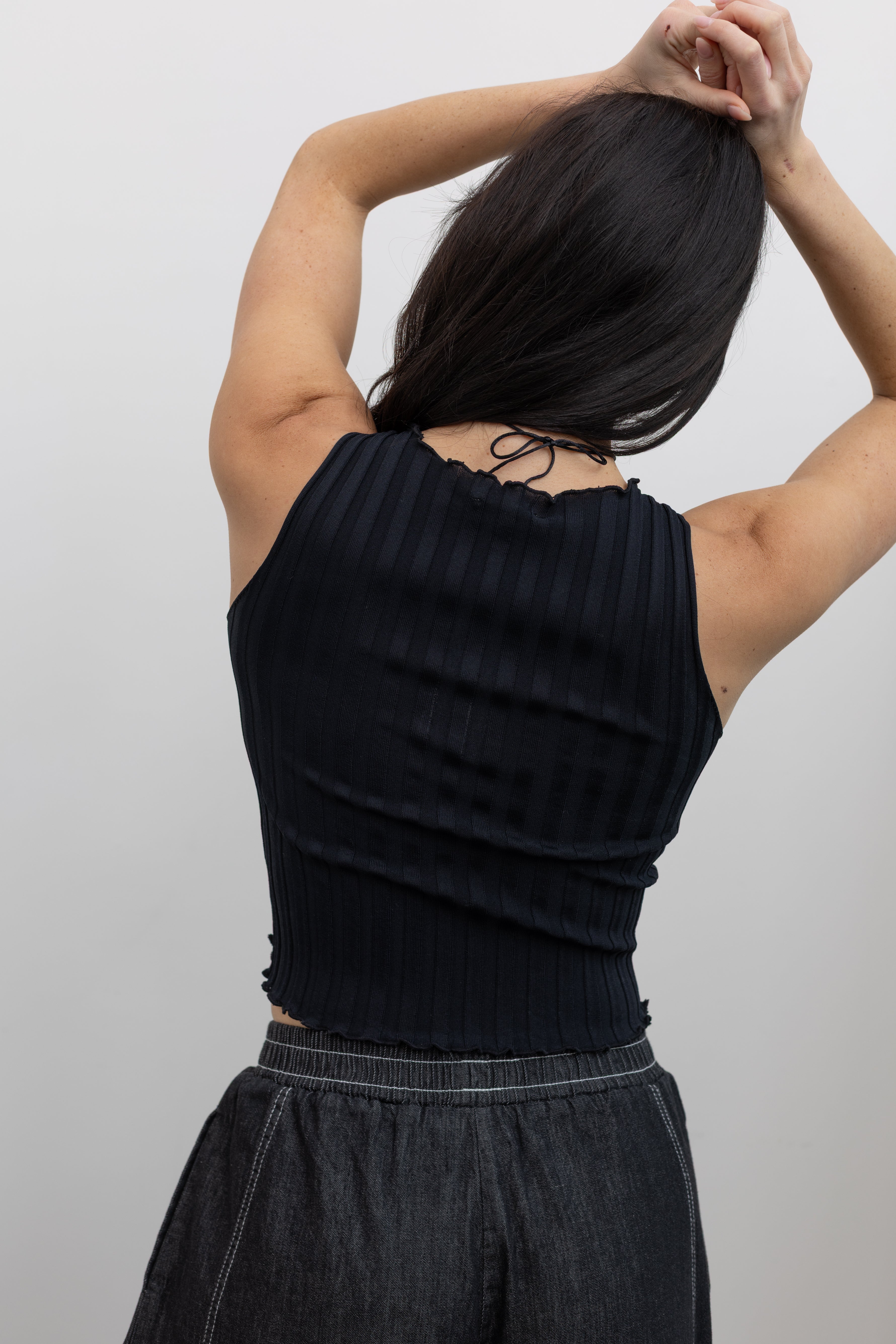 Close-up back view of a woman wearing spring capsule wardrobe essential in a Collective brand black fitted ribbed knit lettuce hem sleeveless top Alas Eius Label black cotton barrel leg pull on pants at an Alas Eius store.