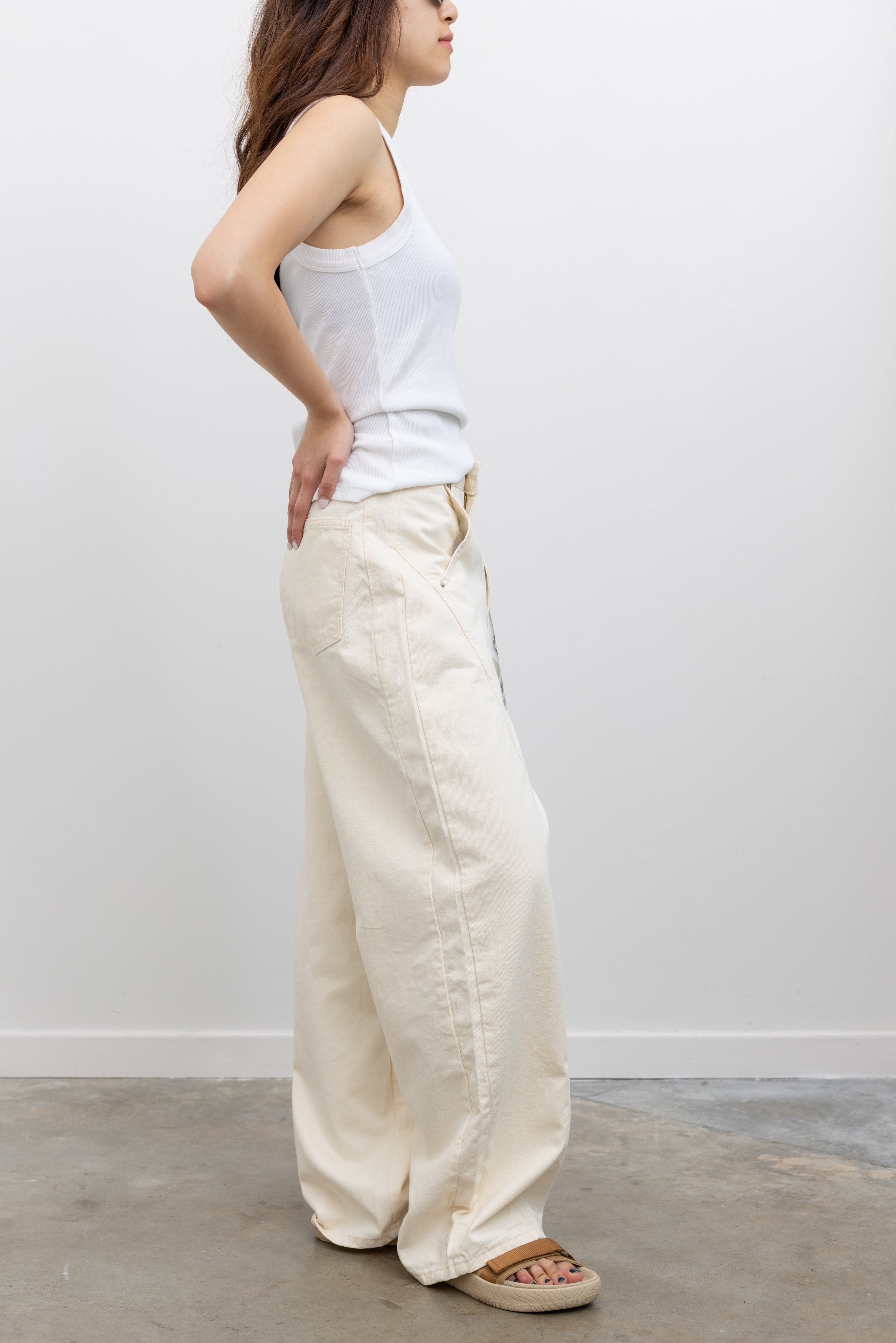 Model wearing minimalist summer fashion in Collective ivory heavy-weight cotton denim jeans with a structured wide-leg silhouette, curved paneling, and relaxed oversized fit paired with an Alas Eius label logo tank and Veja slide sandals at Alas Eius.