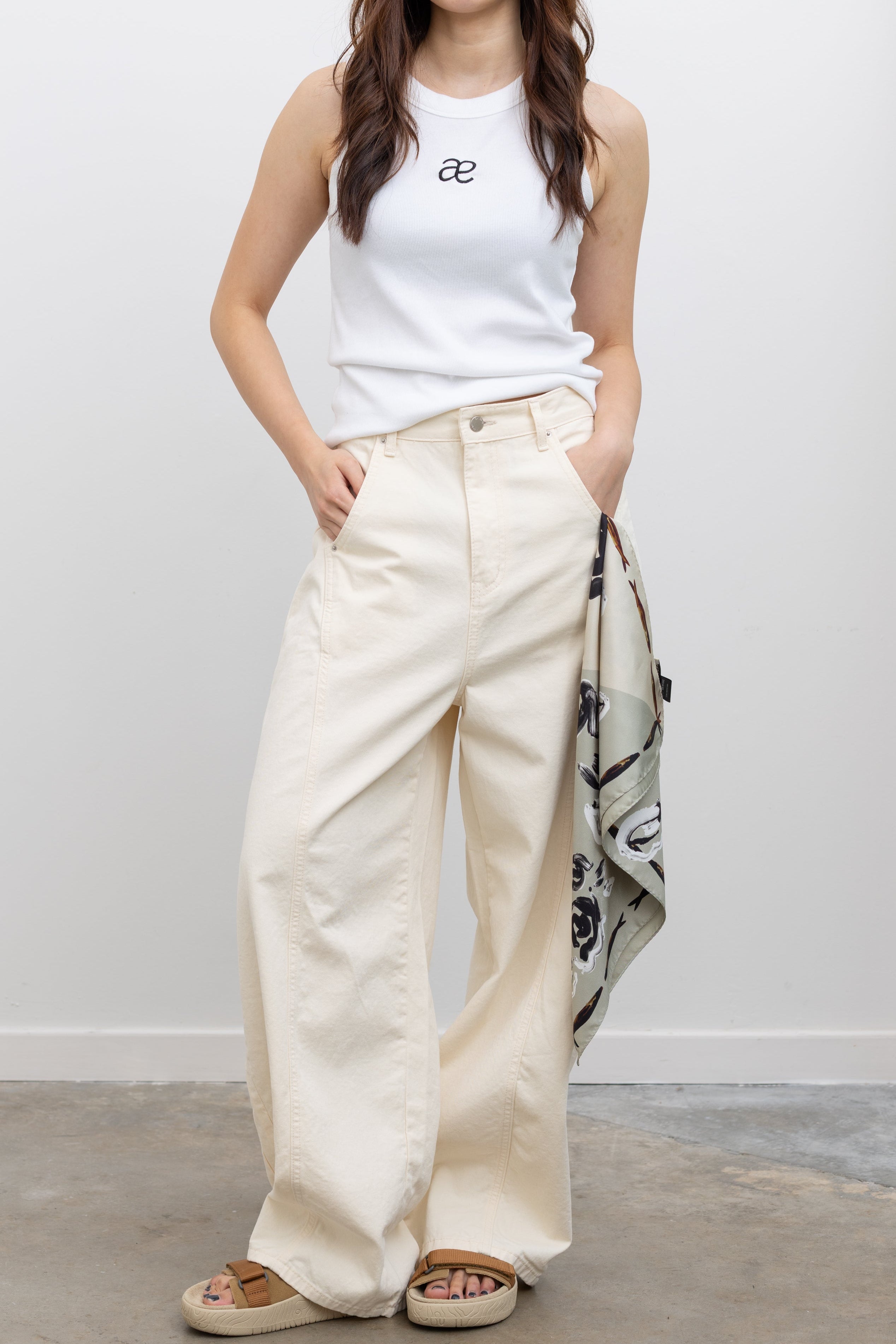 Model wearing Collective cream heavy-weight cotton denim jeans with a structured wide-leg silhouette, curved paneling, and relaxed oversized fit paired with an Alas Eius label logo tank and Veja slide sandals at Alas Eius. 