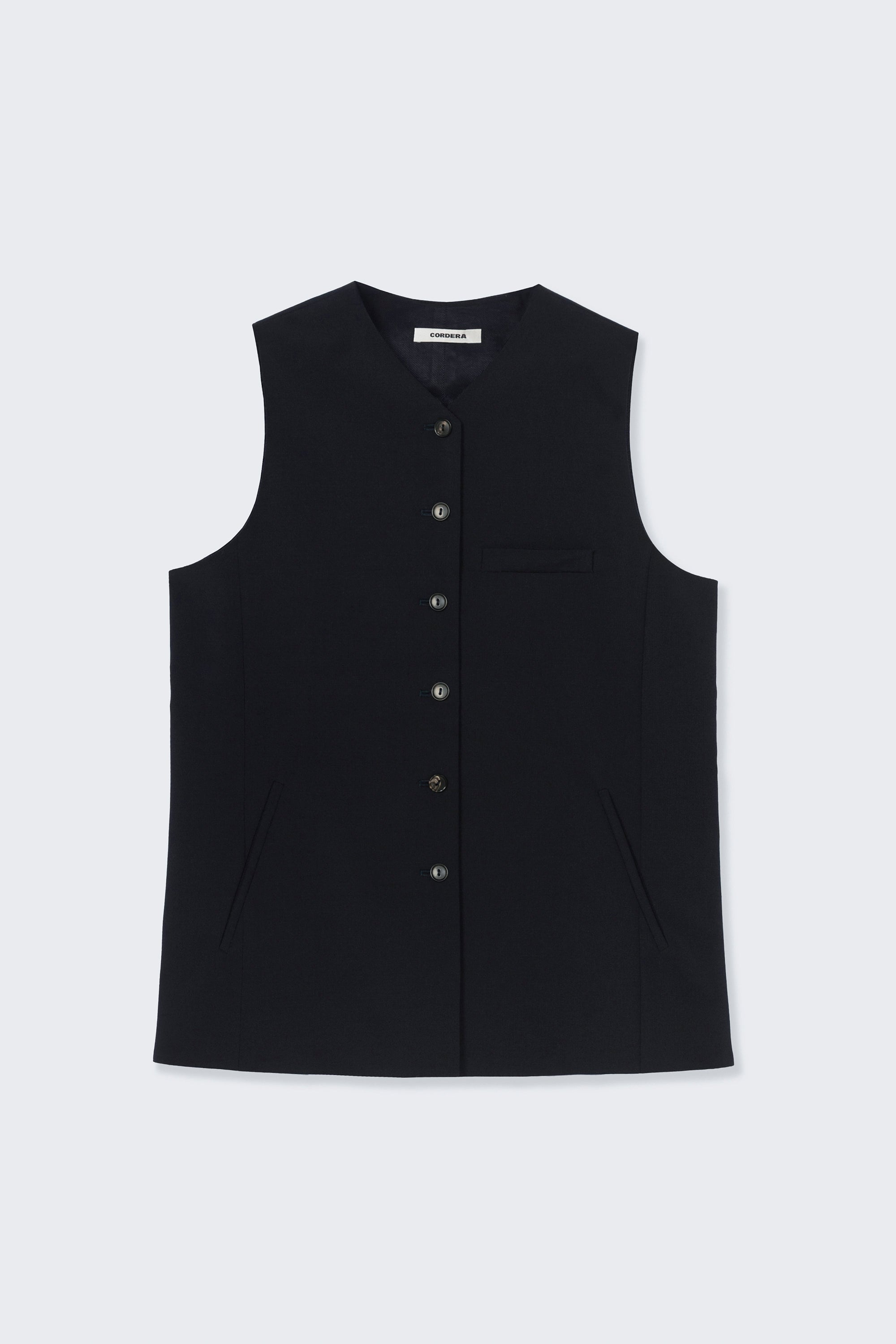 Front view of modern minimalist spring fashion in the Cordera navy blue tailored vest featuring a front button closure and a sharp V-neck collar. Designed with a decorative false pocket and two sleek jetted side pockets at an Alas Eius store. 