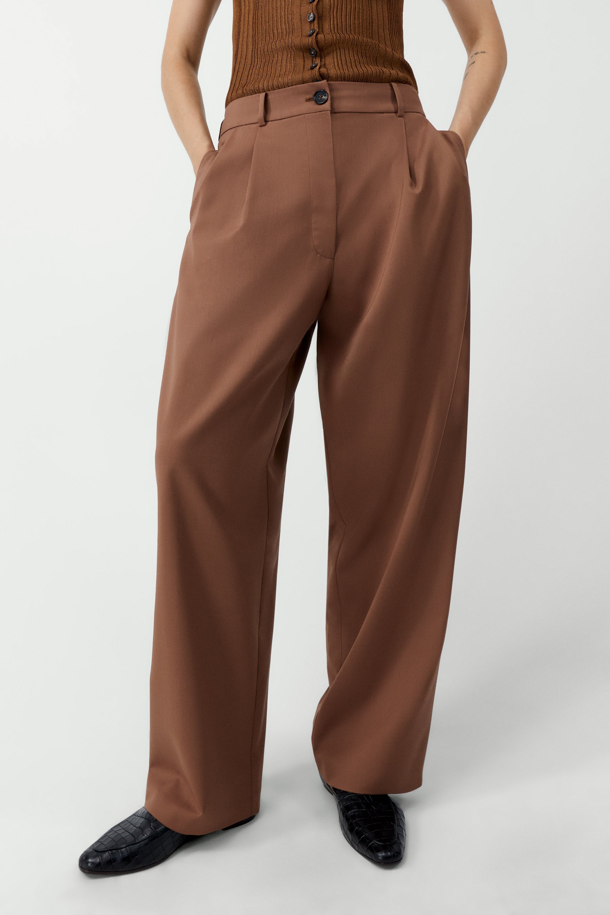 Front view of a woman wearing an elevated wardrobe staple in the Cordera Clove Tailored Masculine Pants featuring a loose, masculine fit with a mid-rise and double front pleats for added structure. Details include jetted side pockets, back welt pockets, an elasticated waistband at the back, zip and button closure, and belt loops at Alas Eius. 