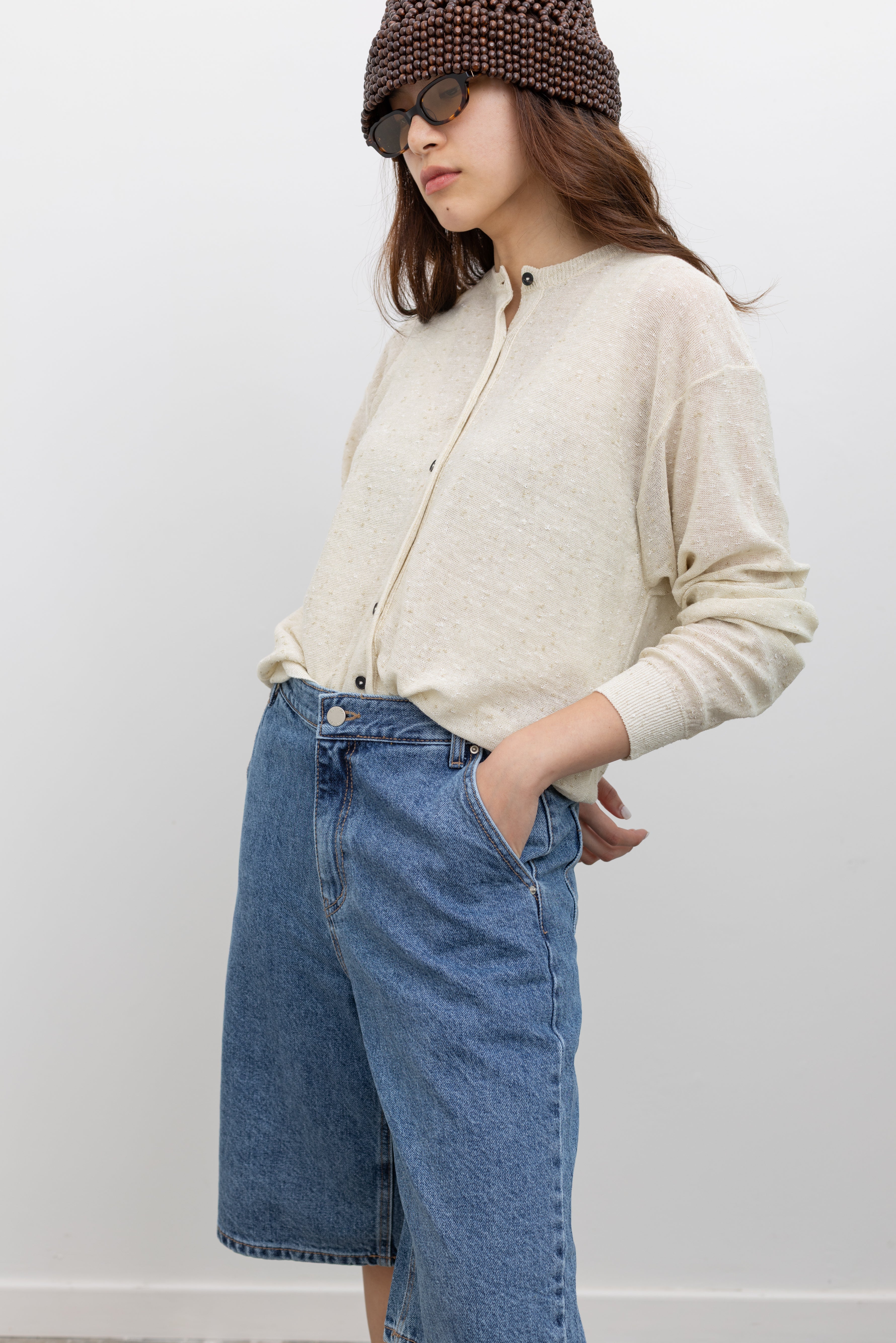 Full-length view of a woman wearing a Cordera cream oversized knit cardigan with front buttons, ribbed trims, and a relaxed silhouette in a flecked linen and cotton blend paired with Collective stone-washed bermuda shorts with a mid-to-high rise, relaxed fit, wide-leg silhouette, and knee-length design at Alas Eius.