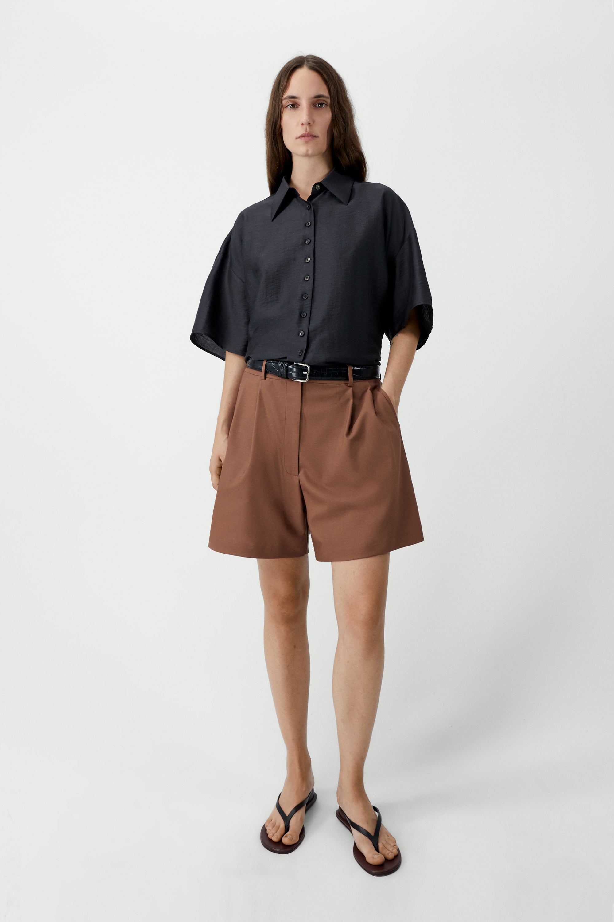Woman standing wearing SS26 contemporary womenswear in the Cordera Espresso Brown Short Sleeve Button Front Shirt featuring an oversized, cocoon-shape, roomy short sleeves, pointy collar, small contrasting buttons down the front at an Alas Eius store. Made in Spain. 
