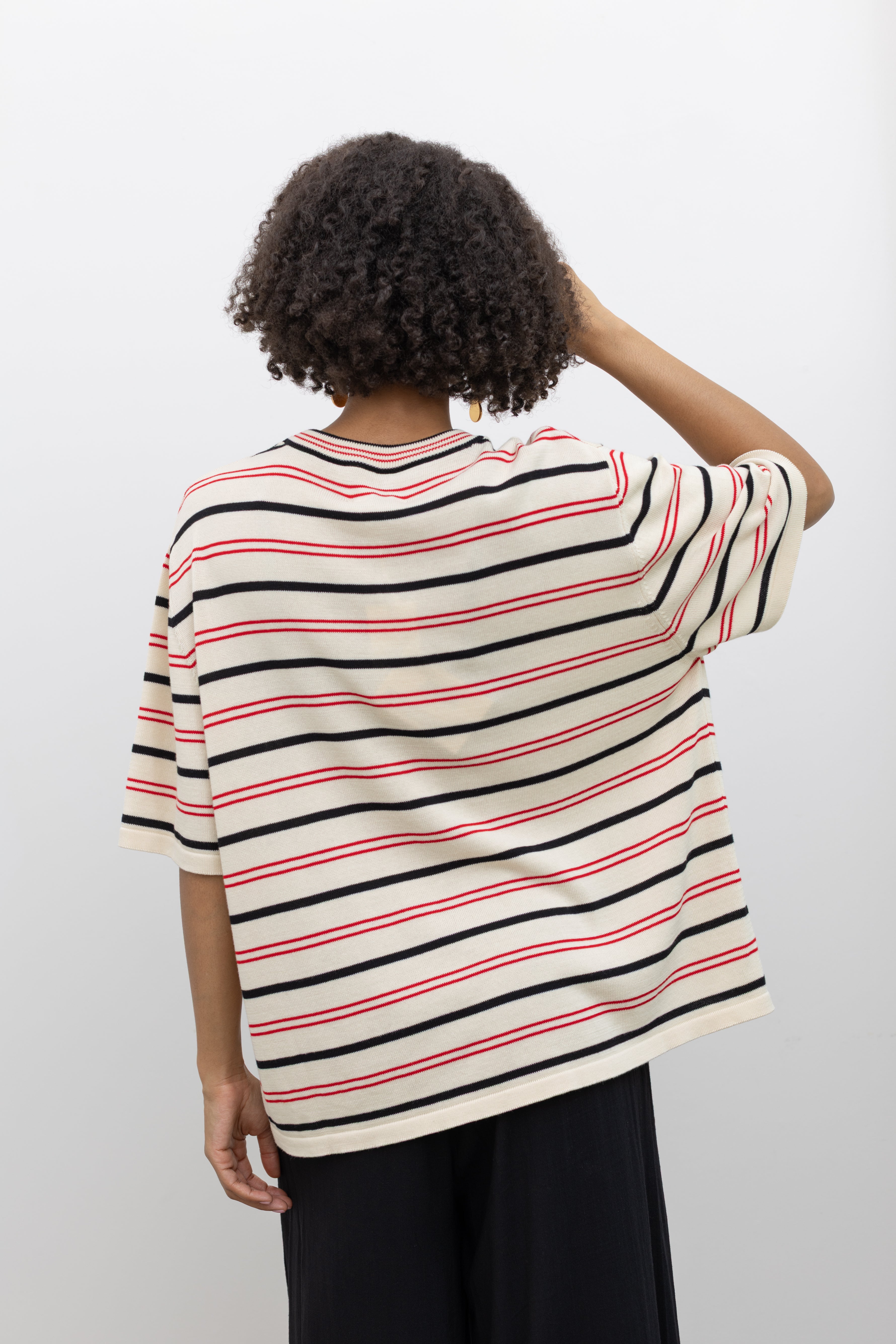 Model wearing modern minimalist summer fashion in a Coreda oversized t-shirt featuring a classic crew neck, red and black stripes in varying widths on an ecru background at Alas Eius.