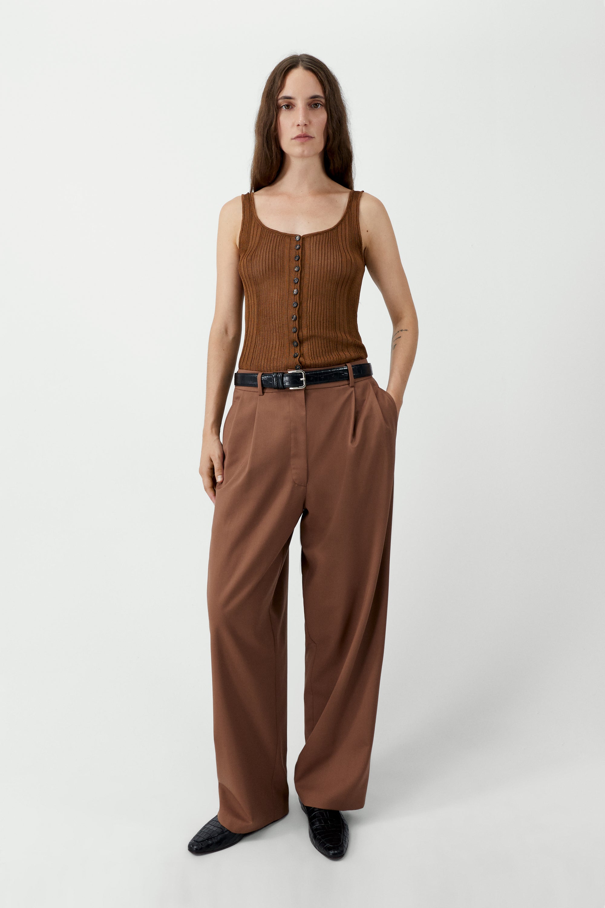 Full length front view of a woman wearing an elevated wardrobe staple in the Cordera Clove Tailored Masculine Pants featuring a loose, masculine fit with a mid-rise and double front pleats for added structure. Details include jetted side pockets, back welt pockets, an elasticated waistband at the back, zip and button closure, and belt loops at Alas Eius. 