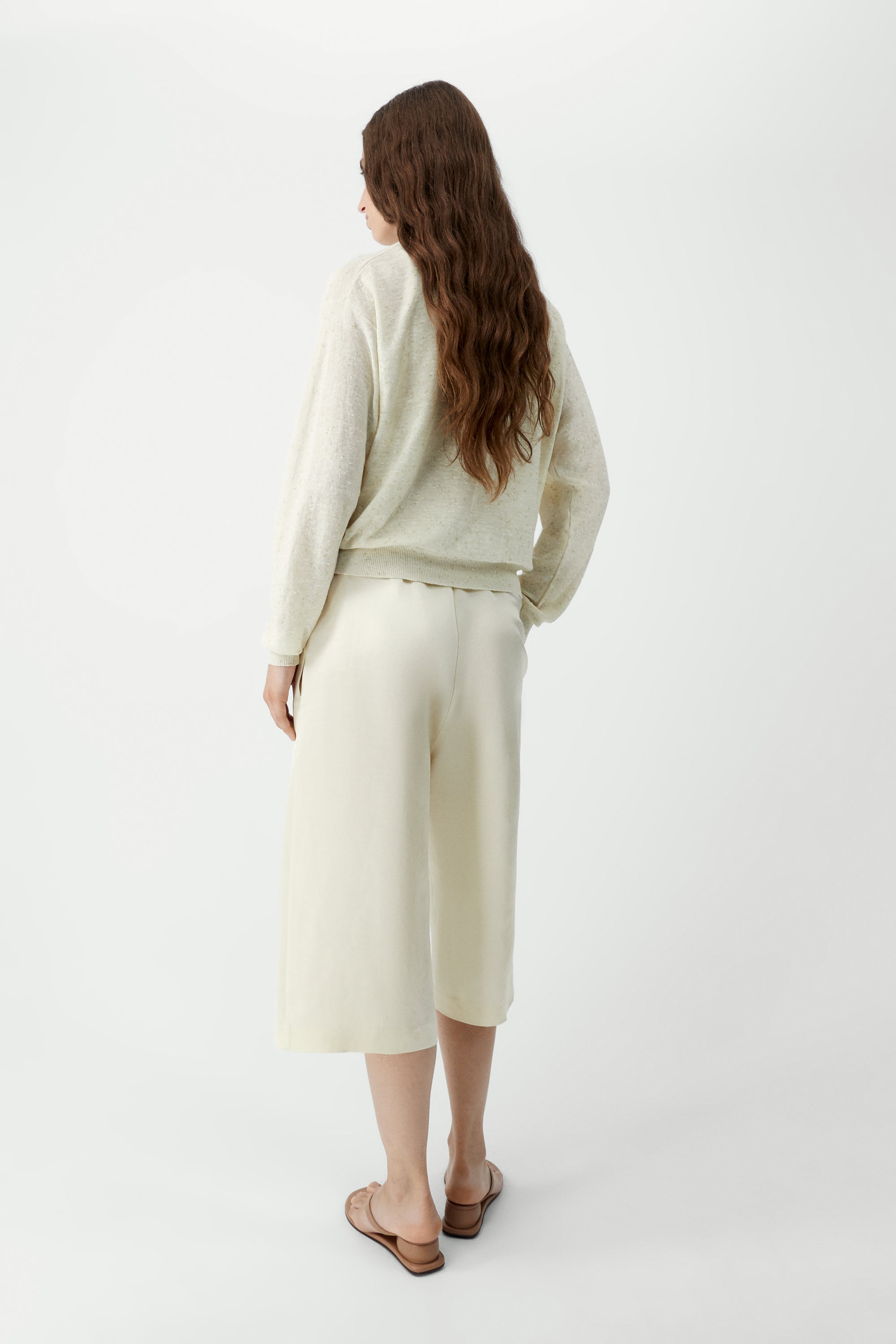 Full length back view of a woman standing wearing an elevated classic wardrobe staple in the Cordera cream linen and cotton blend flecked oversized cardigan featuring a crew neck and finished with ribbed trims at the collar, hem, and cuffs, it fastens with small front buttons at Alas Eius.  

This cardigan is ethically and carefully manufactured in La Coruña, with Japanese knitting machines and finished by hand.