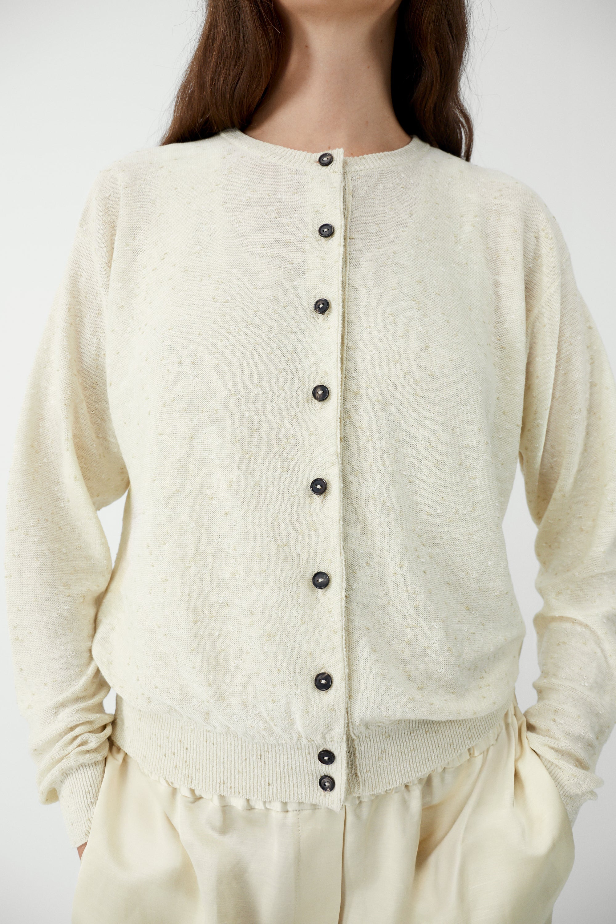 Front view of a woman standing wearing an elevated classic spring fashion staple in the Cordera cream linen and cotton blend flecked oversized cardigan featuring a crew neck and finished with ribbed trims at the collar, hem, and cuffs, it fastens with small front buttons at Alas Eius.  

This cardigan is ethically and carefully manufactured in La Coruña, with Japanese knitting machines and finished by hand.