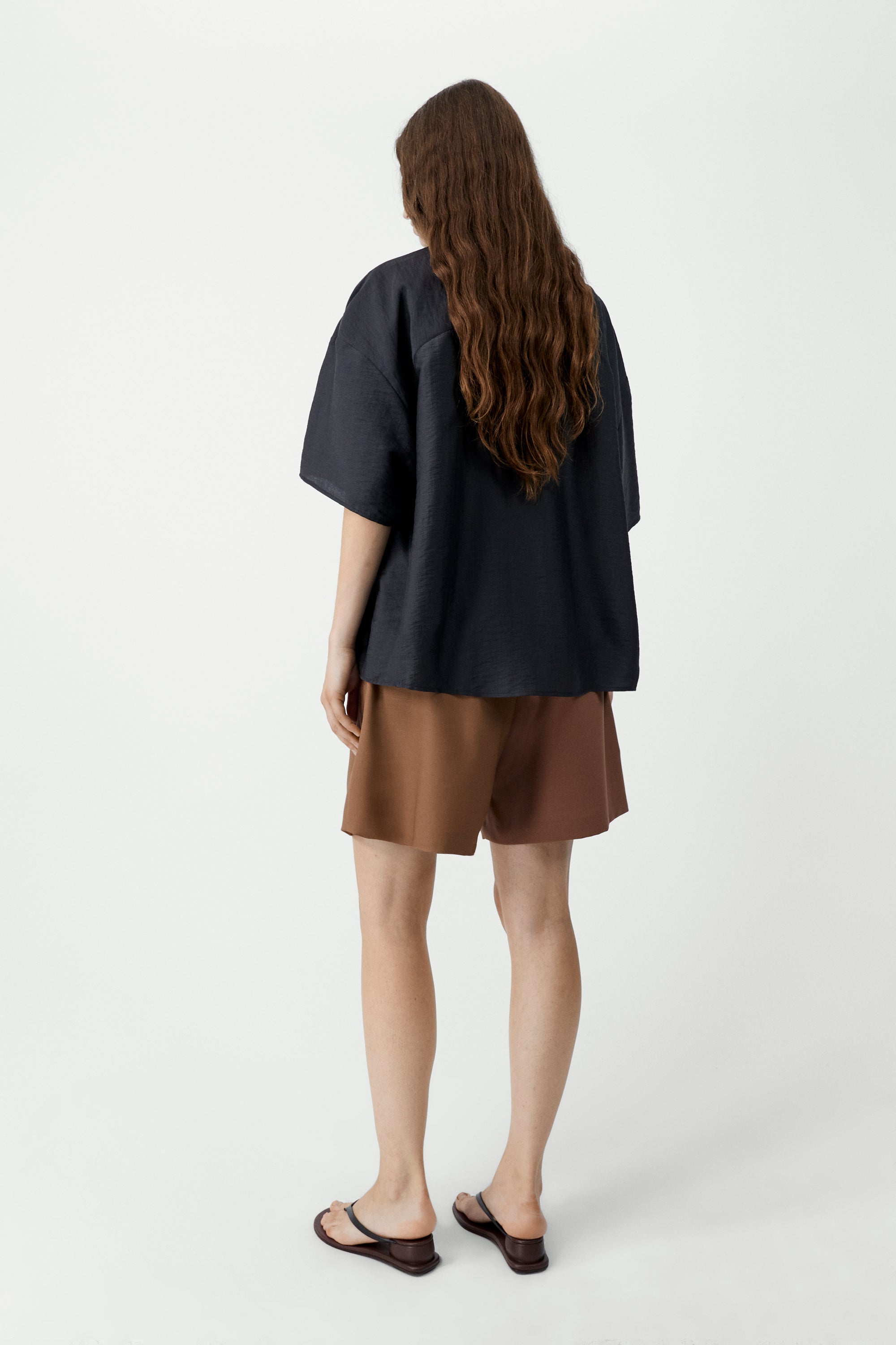 Back view of a woman standing wearing SS26 contemporary womenswear in the Cordera Espresso Brown Short Sleeve Button Front Shirt featuring an oversized, cocoon-shape, roomy short sleeves, pointy collar, small contrasting buttons down the front at an Alas Eius store. Made in Spain. 