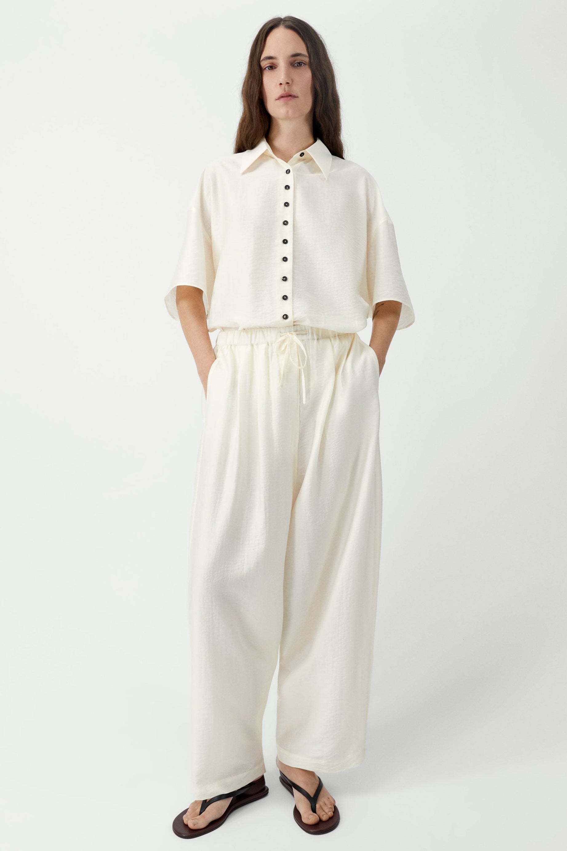 Full length front view of a woman standing wearing spring fashion in Cordera SS26 Marfil maxi pants designed with a fluid silhouette and a relaxed fit. They feature a drawstring elastic waistband and side pockets at an Alas Eius store. Made in Spain.