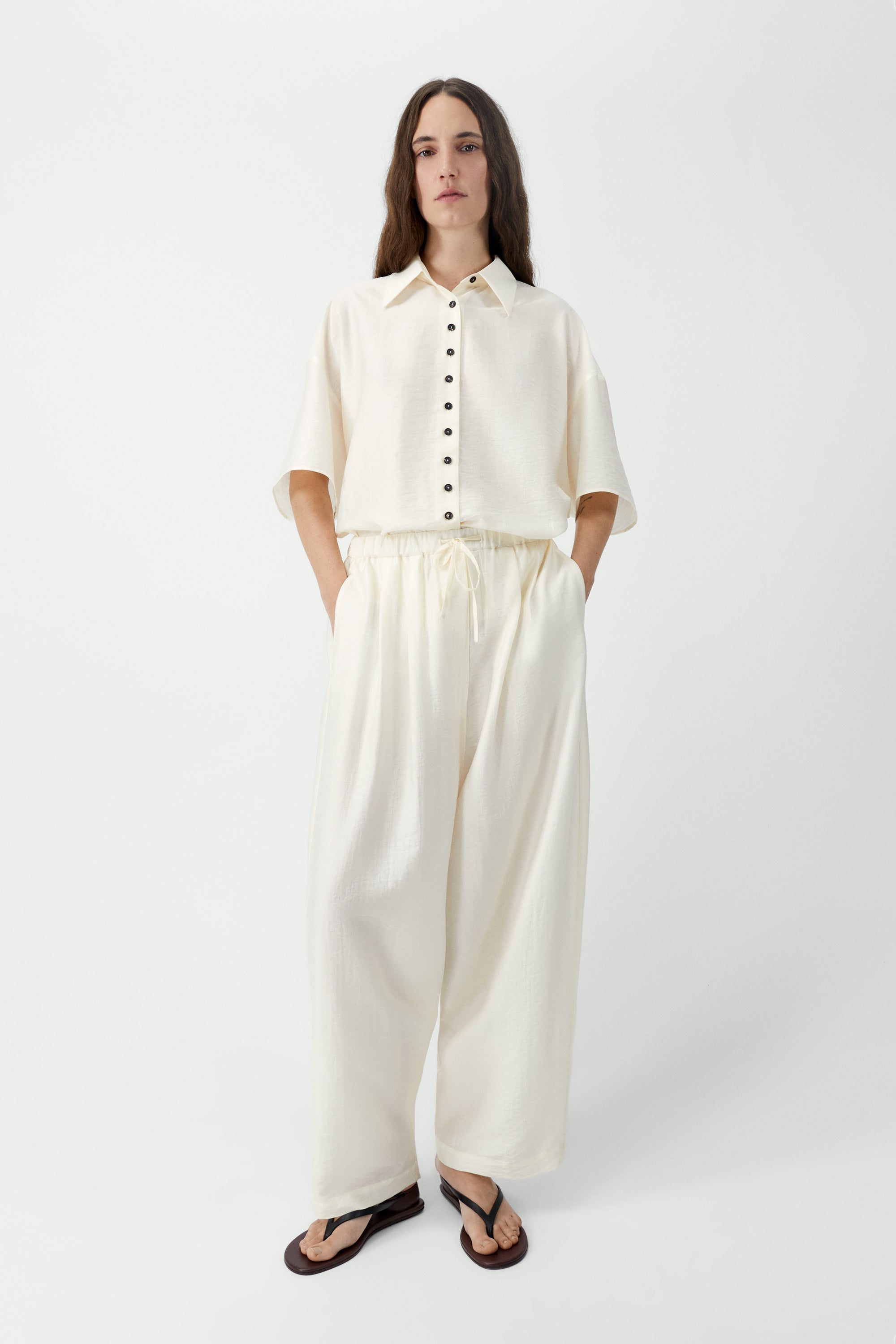 Full length front view of a woman standing wearing spring fashion in Cordera SS26 Marfil maxi pants designed with a fluid silhouette and a relaxed fit. They feature a drawstring elastic waistband and side pockets at an Alas Eius store. Made in Spain.