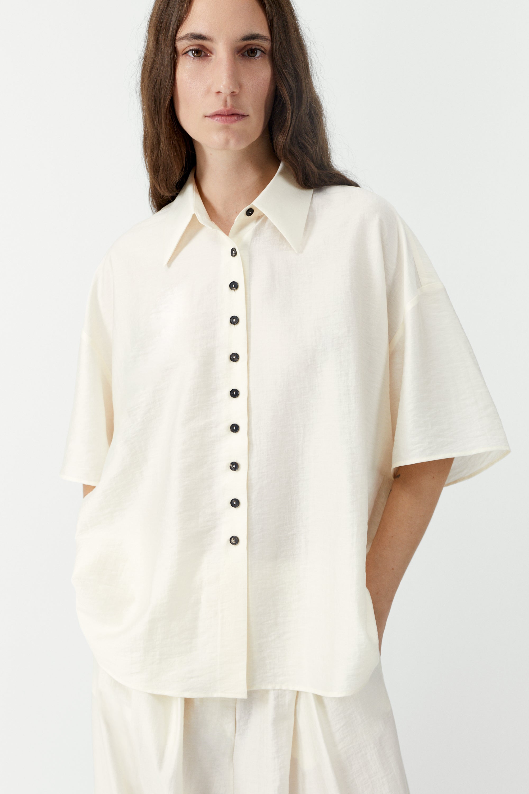 Front view of a woman standing wearing SS26 contemporary womenswear in the Cordera marfil Short Sleeve Button Front Shirt featuring an oversized, cocoon-shape, roomy short sleeves, pointy collar, small contrasting buttons down the front at an Alas Eius store. Made in Spain. 