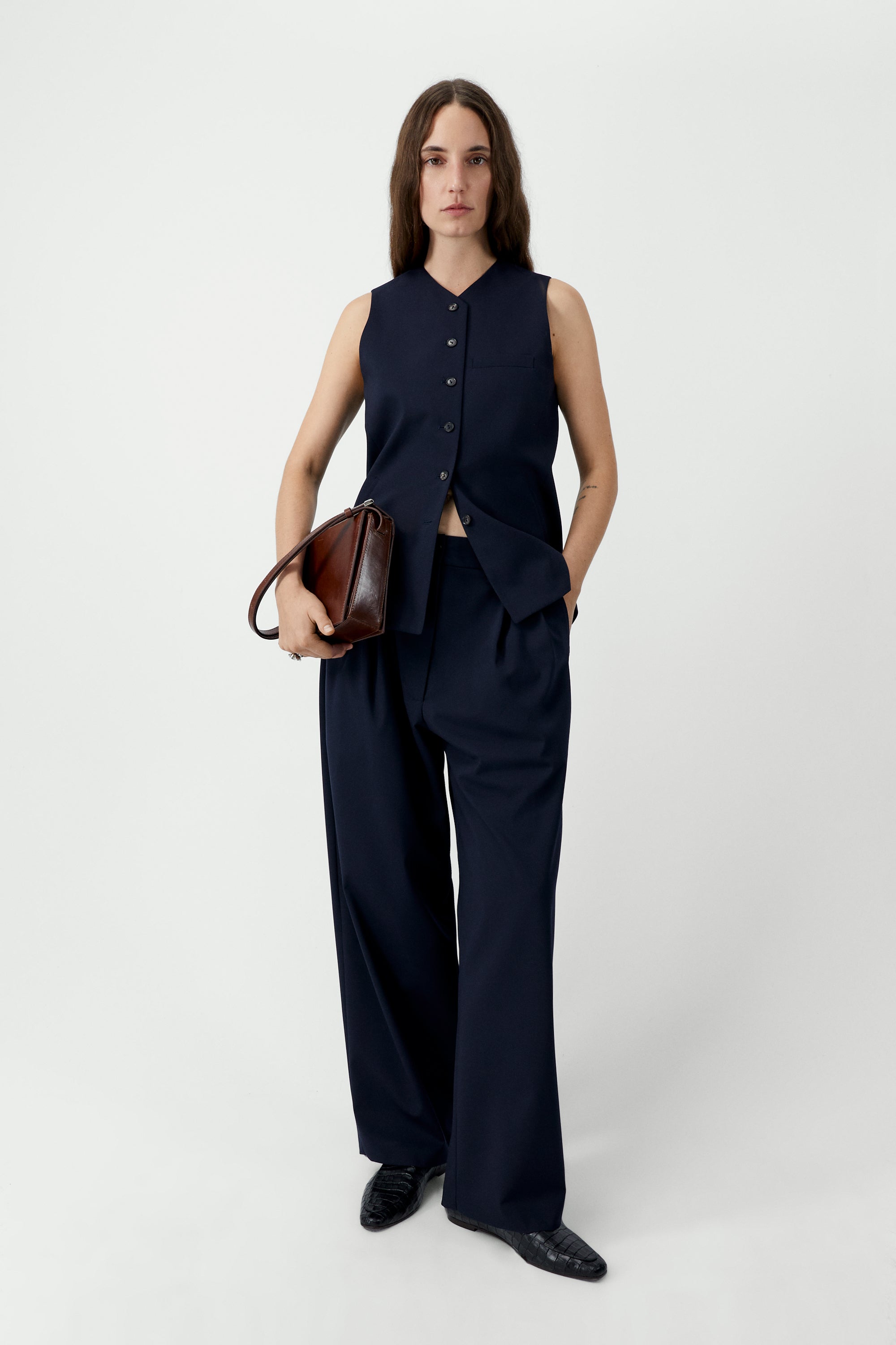 Full length front view of a woman wearing an elevated wardrobe staple in the Cordera Navy Blue Tailored Masculine Pants featuring a loose, masculine fit with a mid-rise and double front pleats for added structure. Details include jetted side pockets, back welt pockets, an elasticated waistband at the back, zip and button closure, and belt loops paired with a Cordera Navy Blue Tailored Vest at Alas Eius. Made in Spain. 