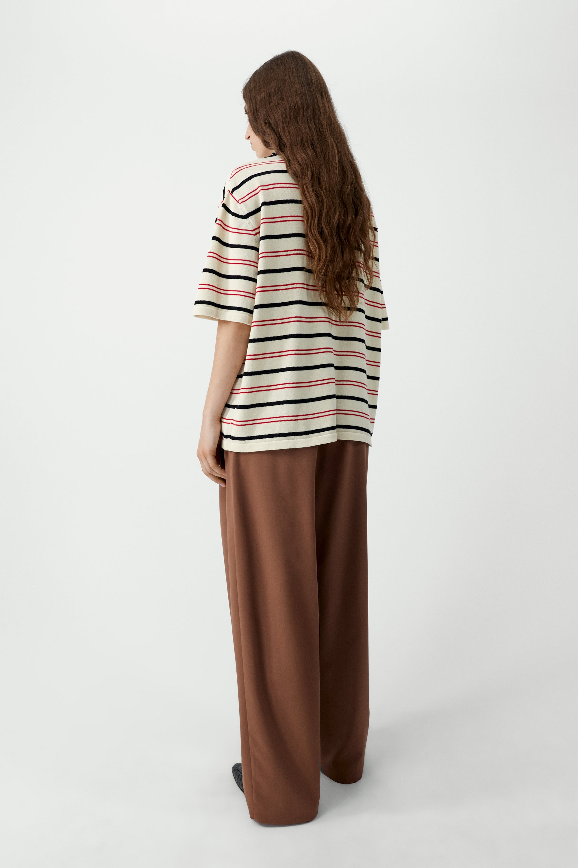 Full length back view of a woman wearing an elevated spring and summer wardrobe essential in a Cordera organic cotton striped oversized t-shirt featuring a classic crew neck and stripes in varying widths, combining bold red and black on an elegant ecru background. Available at Alas Eius. Made in Spain. 