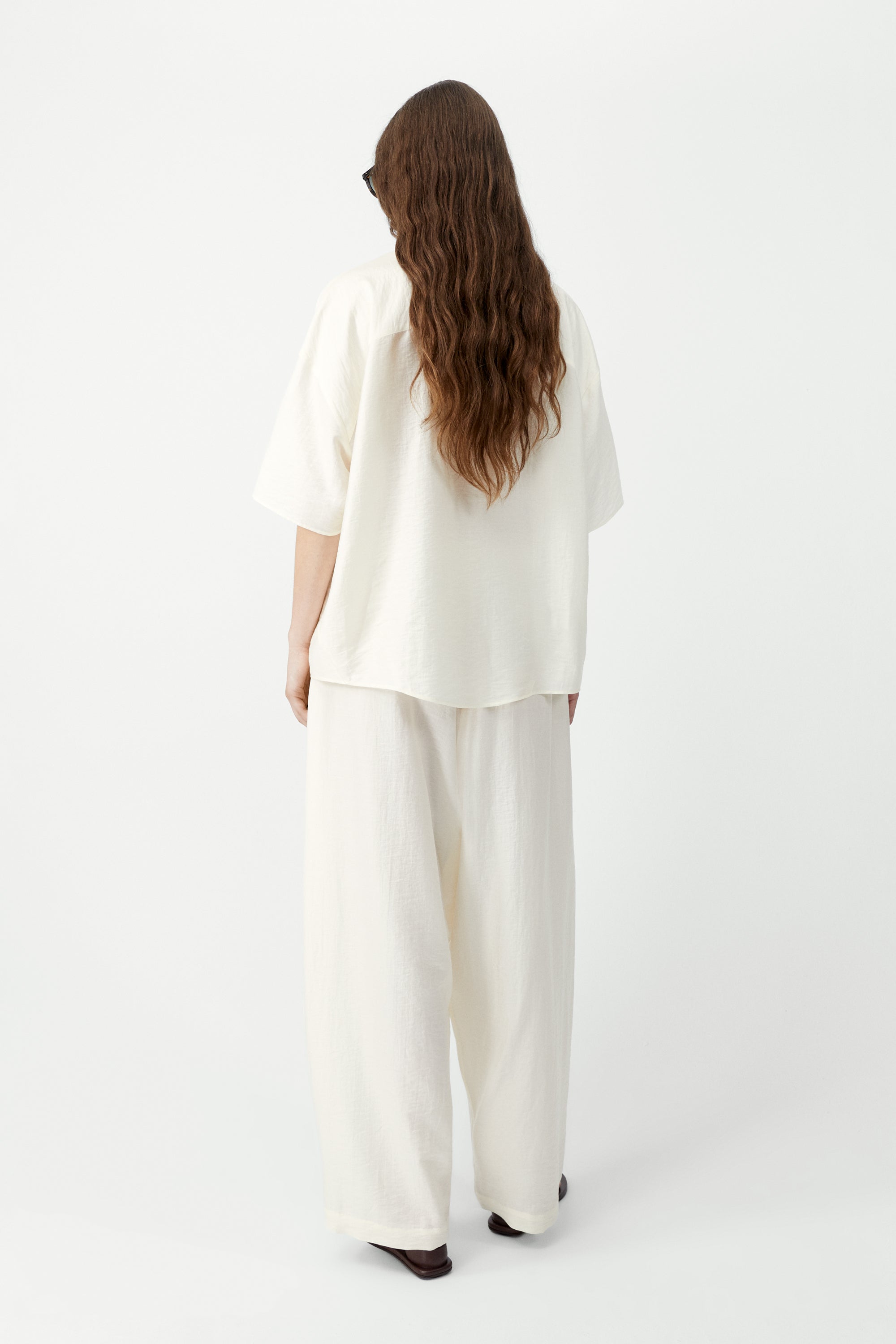 Full length back view of a woman standing wearing SS26 contemporary womenswear in the Cordera marfil Short Sleeve Button Front Shirt featuring an oversized, cocoon-shape, roomy short sleeves, pointy collar, small contrasting buttons down the front at an Alas Eius store. Made in Spain. 