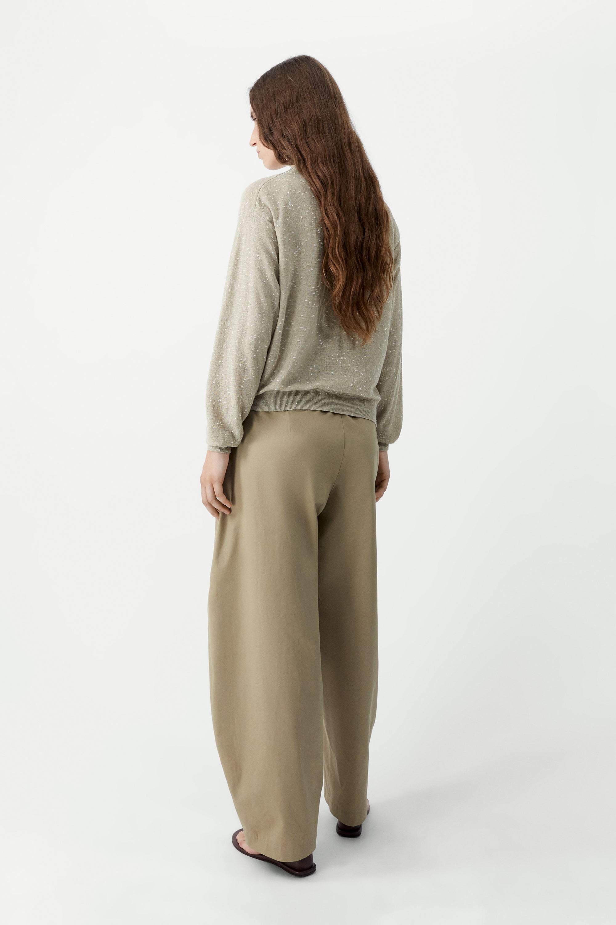 Full length back view of a woman standing wearing an elevated classic wardrobe staple in the Cordera cream linen and cotton blend flecked oversized cardigan featuring a crew neck and finished with ribbed trims at the collar, hem, and cuffs, it fastens with small front buttons at Alas Eius.  

This cardigan is ethically and carefully manufactured in La Coruña, with Japanese knitting machines and finished by hand.