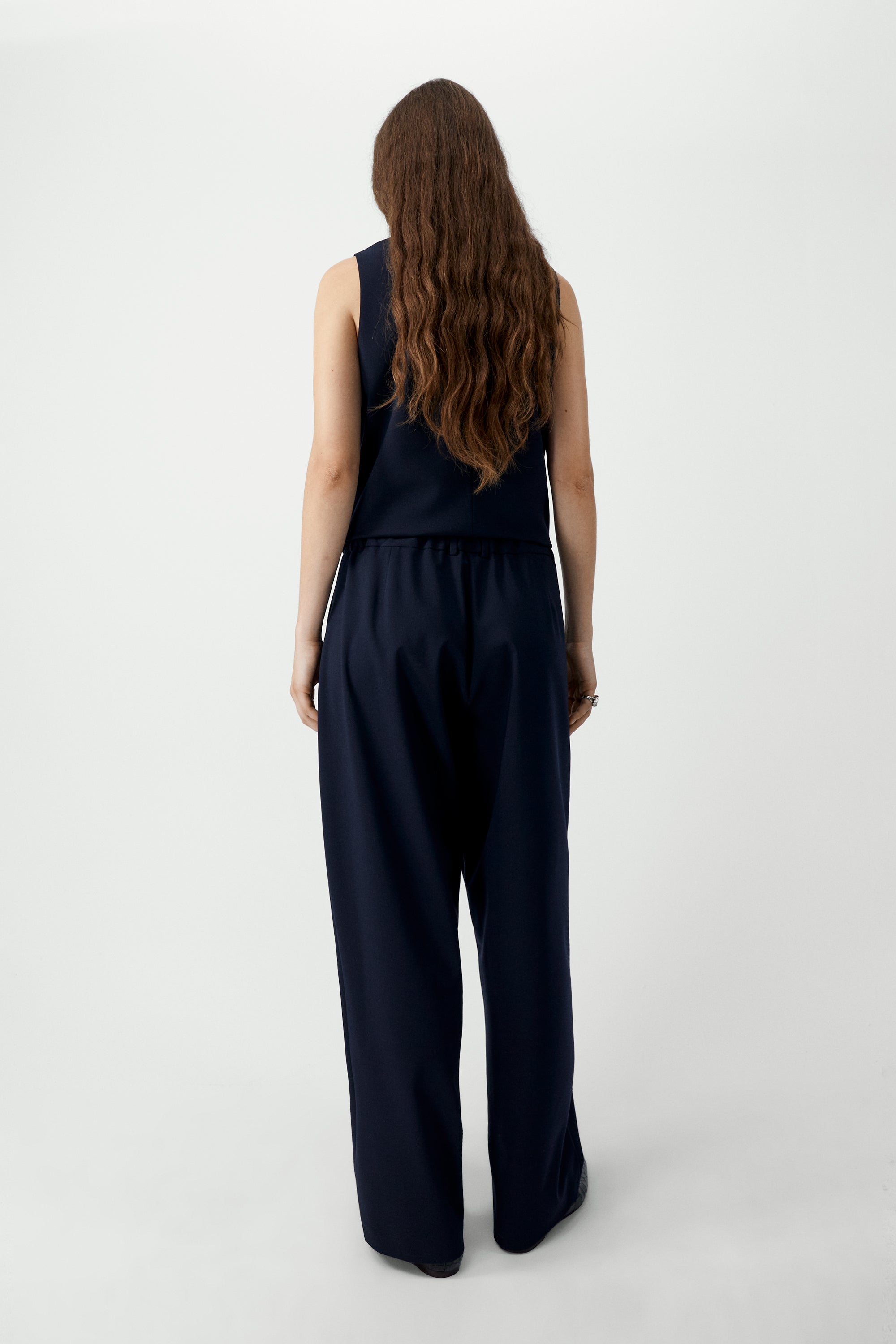 Back view of a woman wearing an elevated wardrobe staple in the Cordera Navy Blue Tailored Masculine Pants featuring a loose, masculine fit with a mid-rise and double front pleats for added structure. Details include jetted side pockets, back welt pockets, an elasticated waistband at the back, zip and button closure, and belt loops paired with a Cordera Navy Blue Tailored Vest at Alas Eius. Made in Spain. 