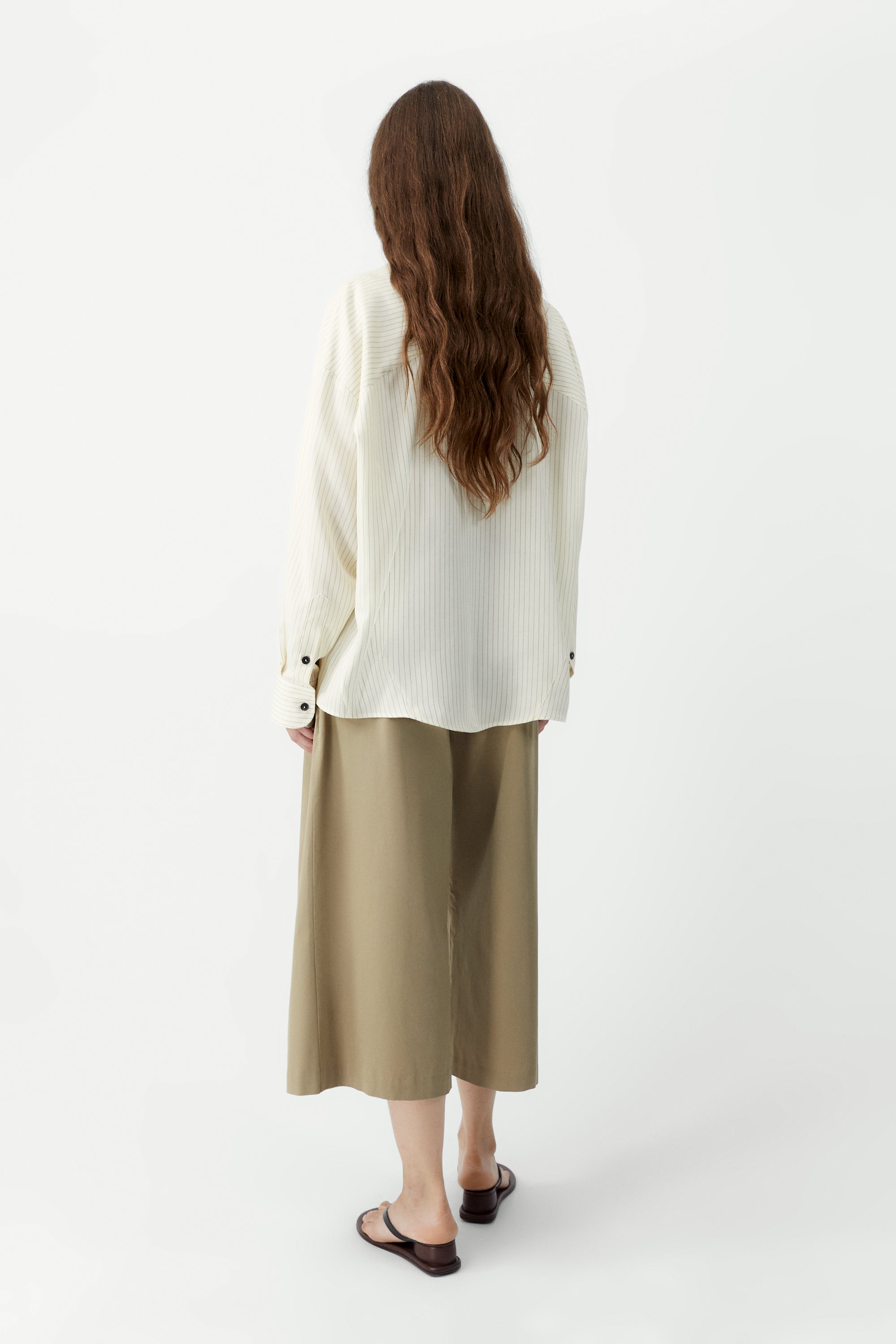 Full length back view of a woman standing wearing a classic spring fashion staple in the Cordera Cream Oversized Pencil Striped Shirt featuring an Italian collar and a front button closure at Alas Eius. 