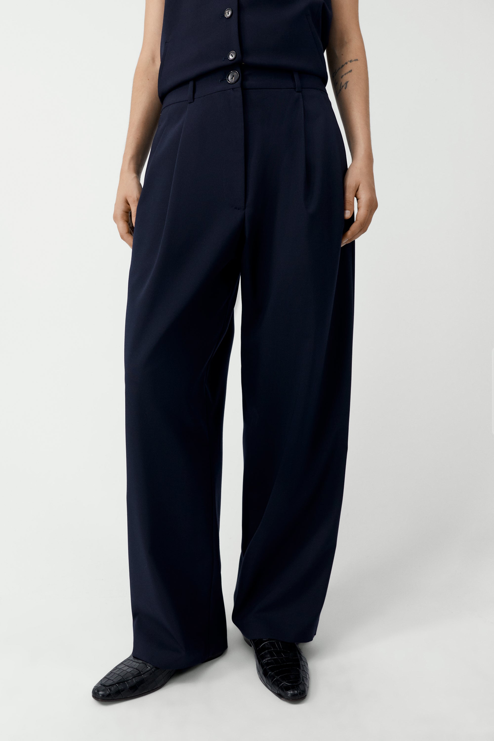 Front view of a woman wearing an elevated wardrobe staple in the Cordera Navy Blue Tailored Masculine Pants featuring a loose, masculine fit with a mid-rise and double front pleats for added structure. Details include jetted side pockets, back welt pockets, an elasticated waistband at the back, zip and button closure, and belt loops paired with a Cordera Navy Blue Tailored Vest at Alas Eius. Made in Spain. 