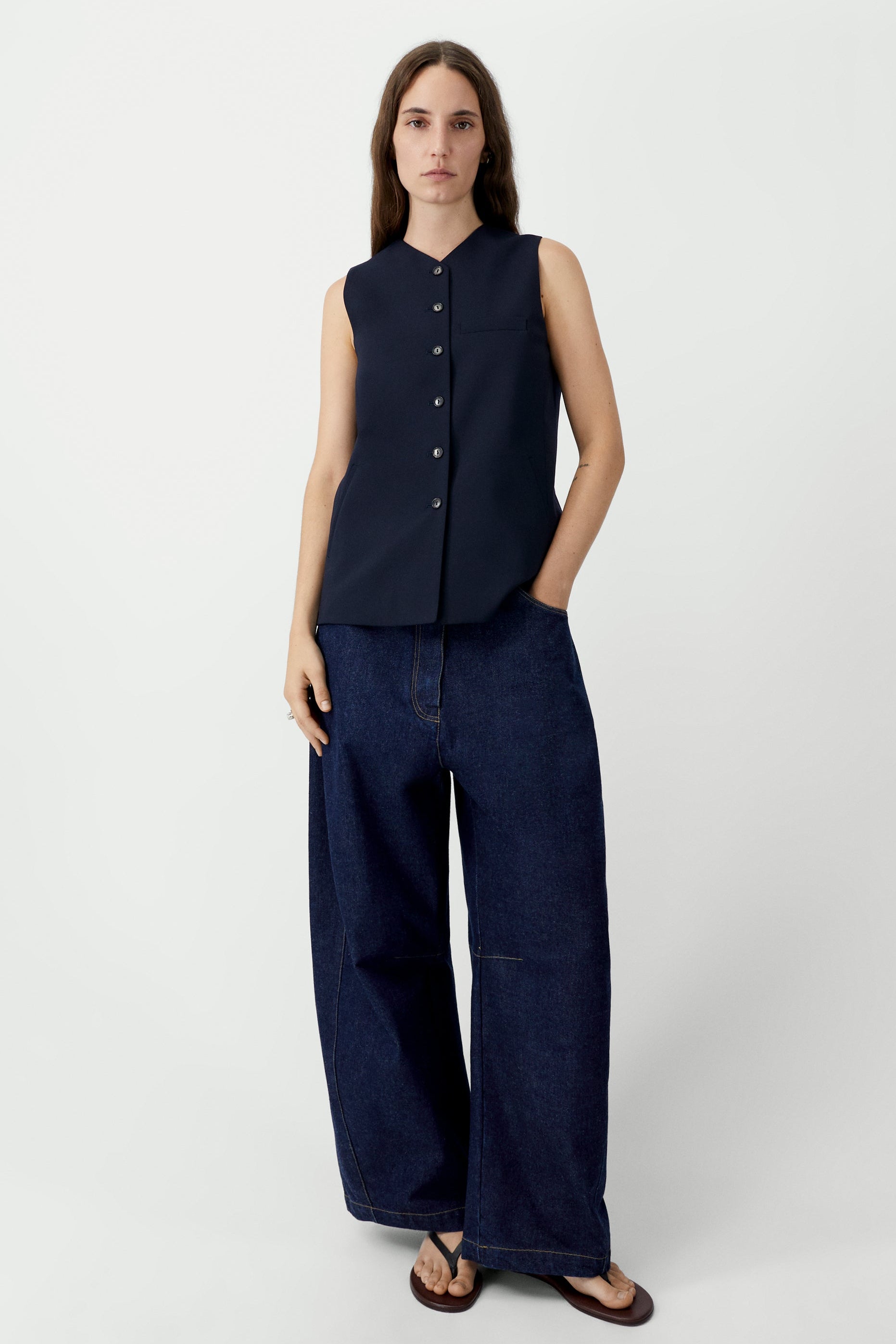 Full length front view of a woman standing wearing modern minimalist spring fashion in the Cordera navy blue tailored vest featuring a front button closure and a sharp V-neck collar. Designed with a decorative false pocket and two sleek jetted side pockets paired with dark wash denim barrel leg jeans and sandals at an Alas Eius store. 