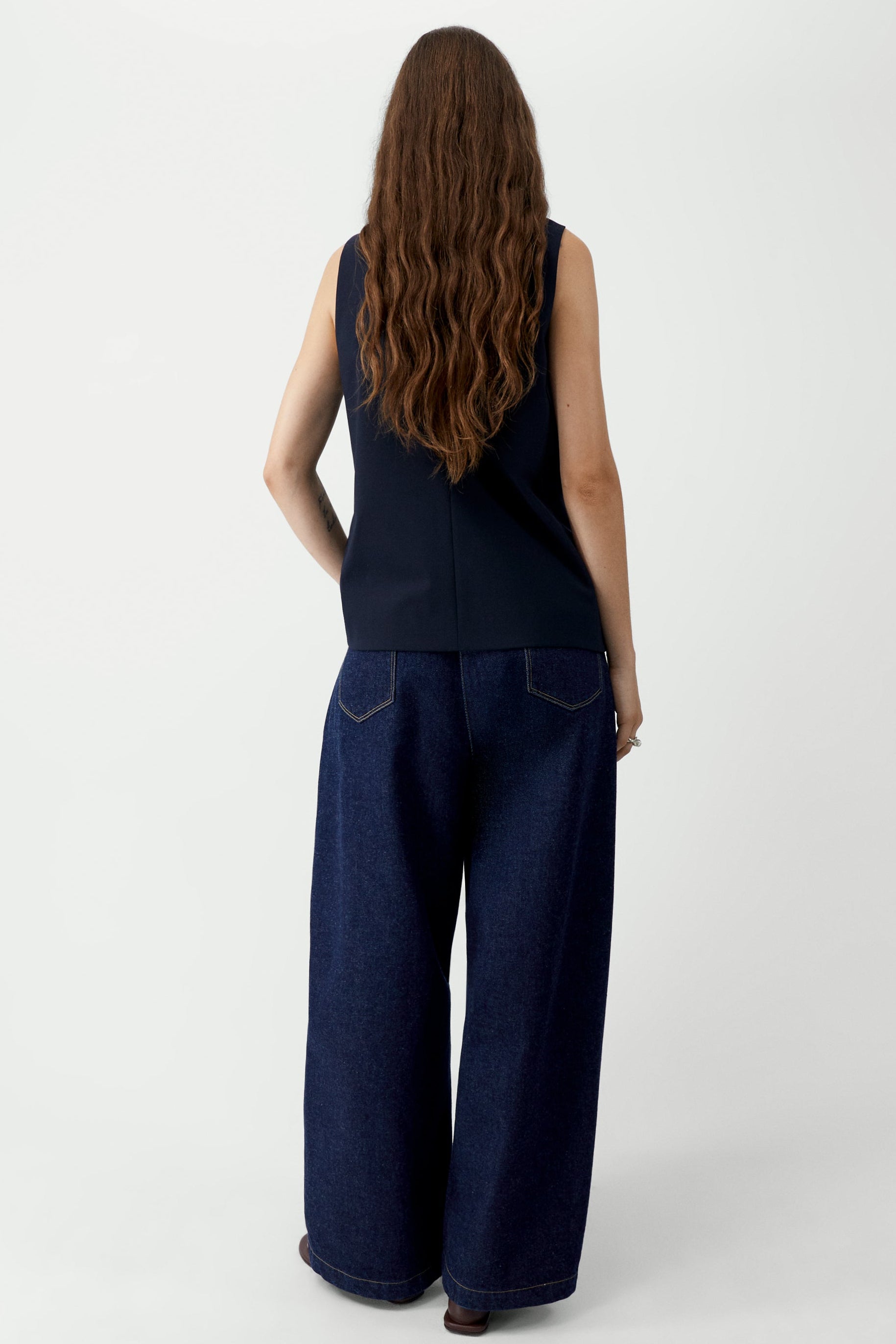 Full length back view of a woman standing wearing modern minimalist spring fashion in the Cordera navy blue tailored vest featuring a front button closure and a sharp V-neck collar. Designed with a decorative false pocket and two sleek jetted side pockets paired with dark wash denim barrel leg jeans and sandals at an Alas Eius store. 
