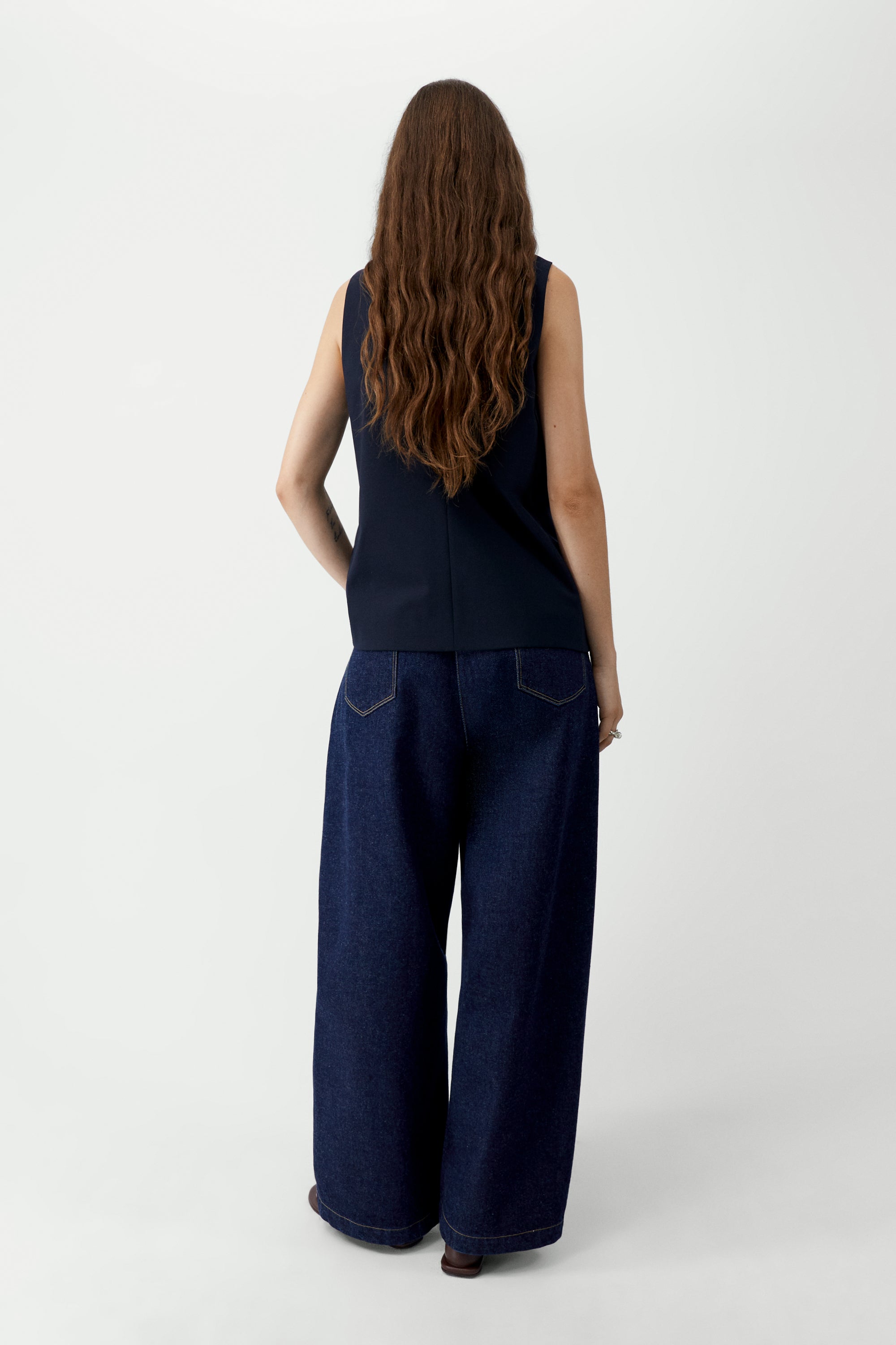 Full length back view of a woman standing wearing modern minimalist spring fashion in the Cordera navy blue tailored vest featuring a front button closure and a sharp V-neck collar. Designed with a decorative false pocket and two sleek jetted side pockets paired with dark wash denim barrel leg jeans and sandals at an Alas Eius store. 