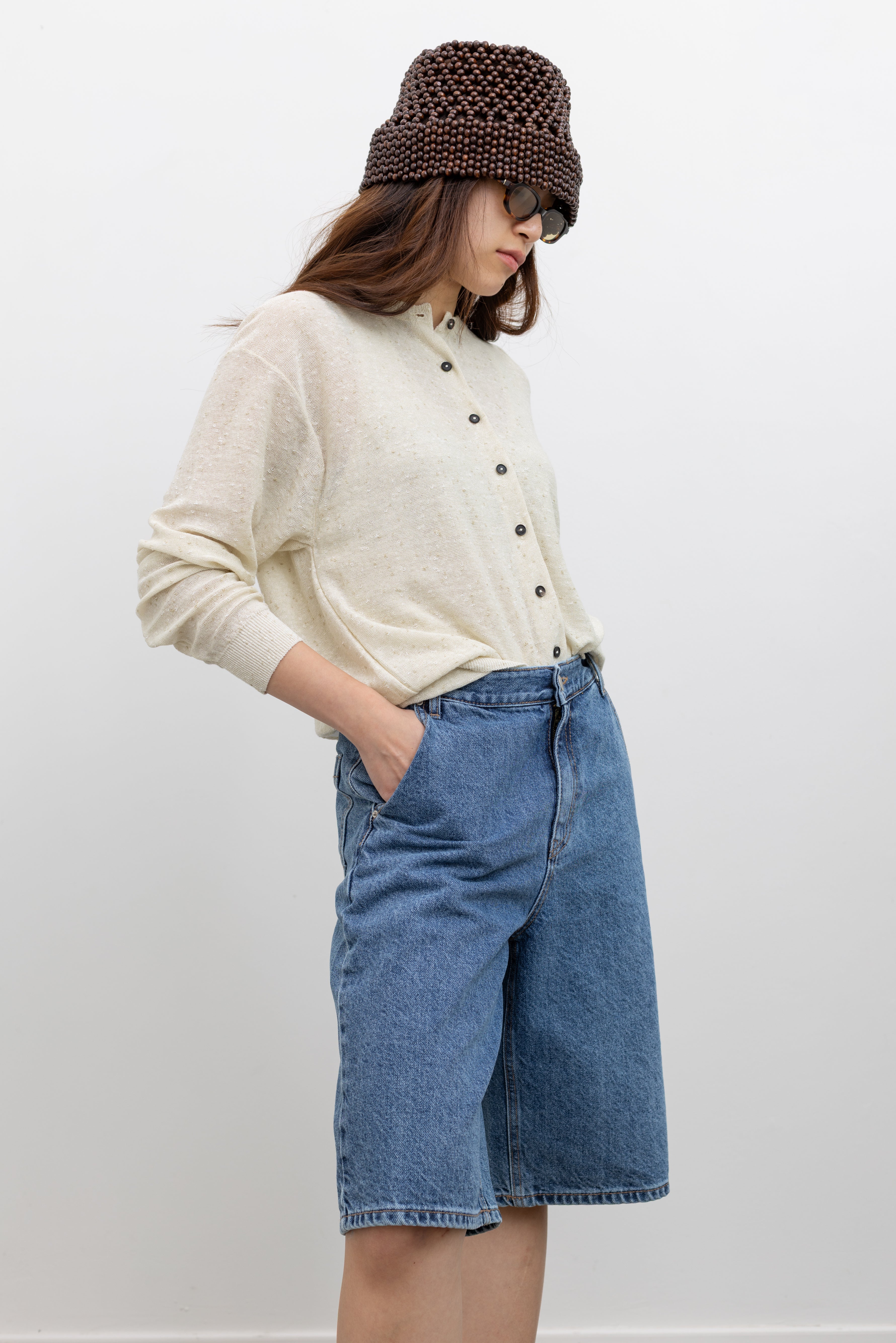 Model wearing a Cordera cream knit cardigan with front buttons and a relaxed silhouette in a flecked linen and cotton blend paired with Collective stone-washed light blue denim bermuda shorts with a wide-leg, and knee-length design paired with a Cordera brown wooden beaded bucket hat at Alas Eius.