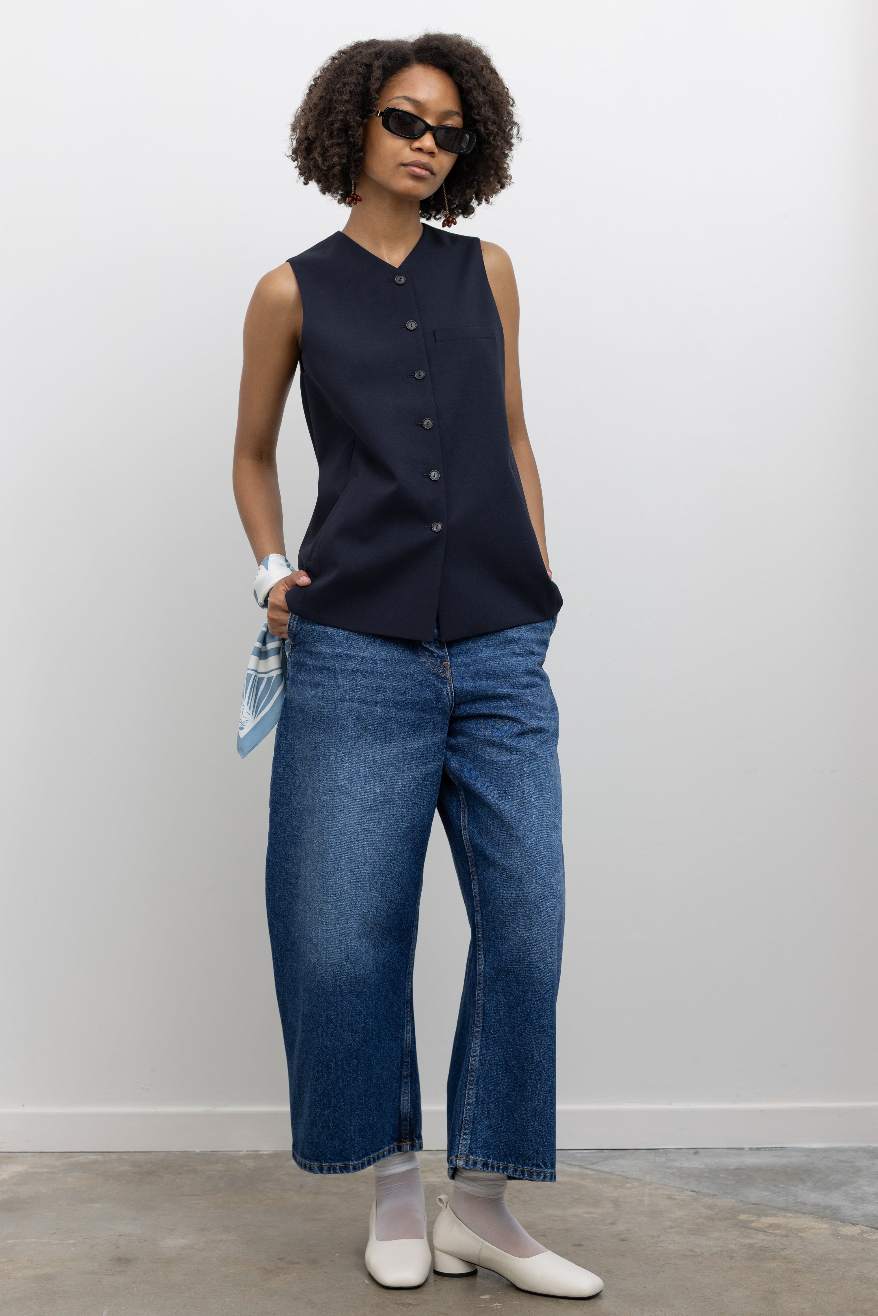Full-length view of a woman styled in a navy tailored vest with V-neck and button front, paired with medium wash cropped barrel-leg denim jeans and white leather pumps at Alas Eius.