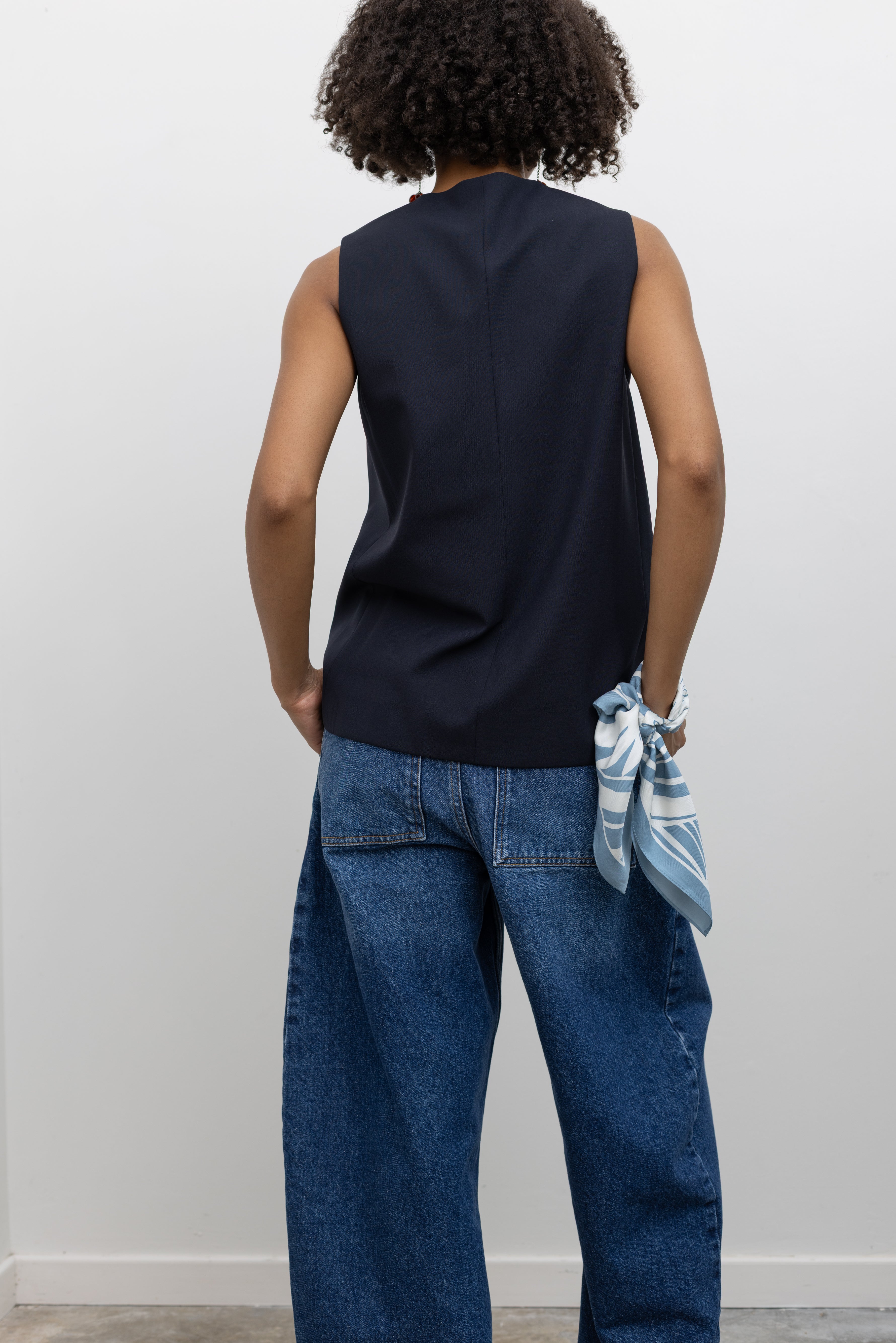 Back view of a woman styled in a Cordera navy blue tailored vest with V-neck and button front, paired with medium wash barrel-leg denim jeans at an Alas Eius store. 