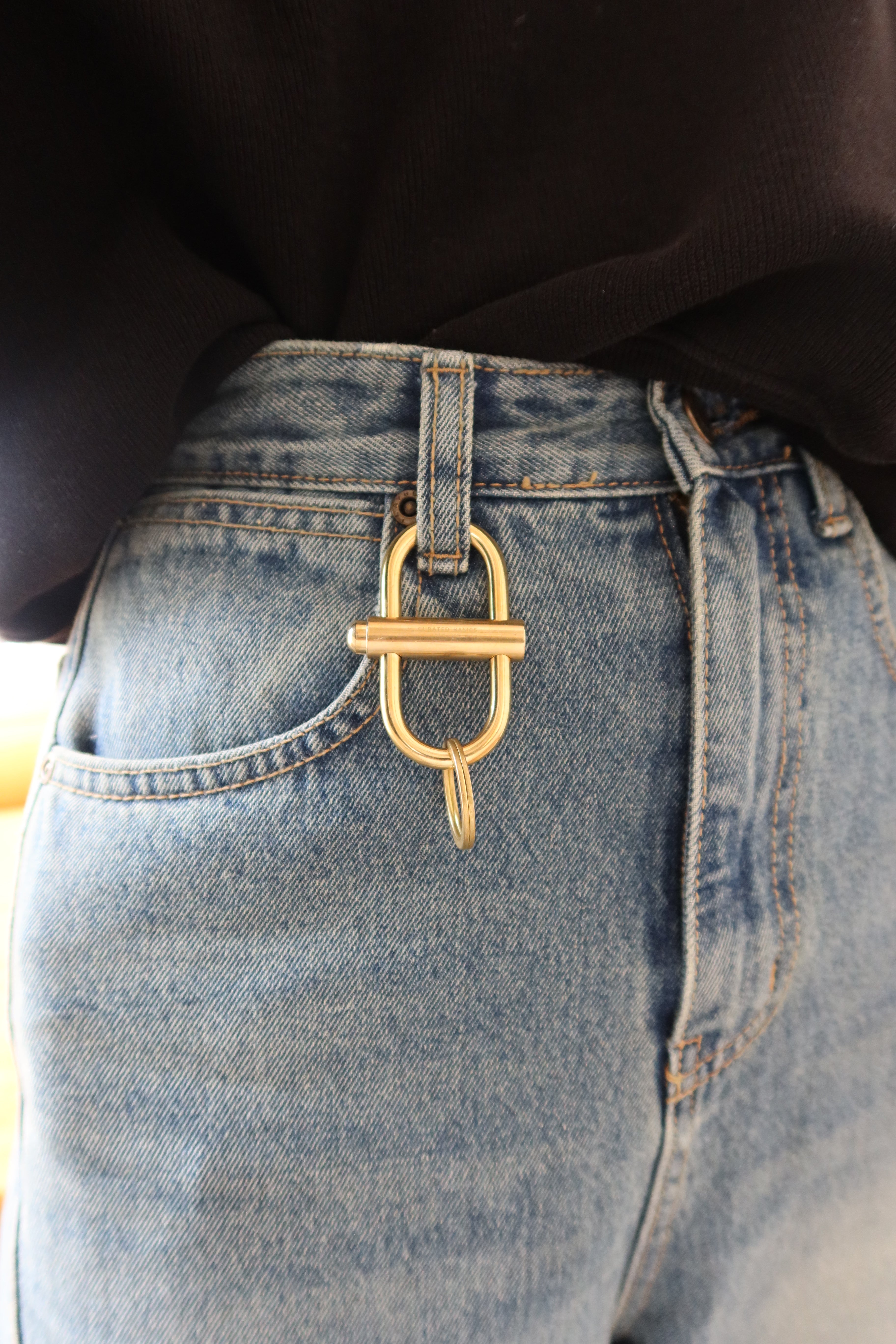 A Curated Basics brand oval brass lock keychain on a belt loop at Alas Eius. 