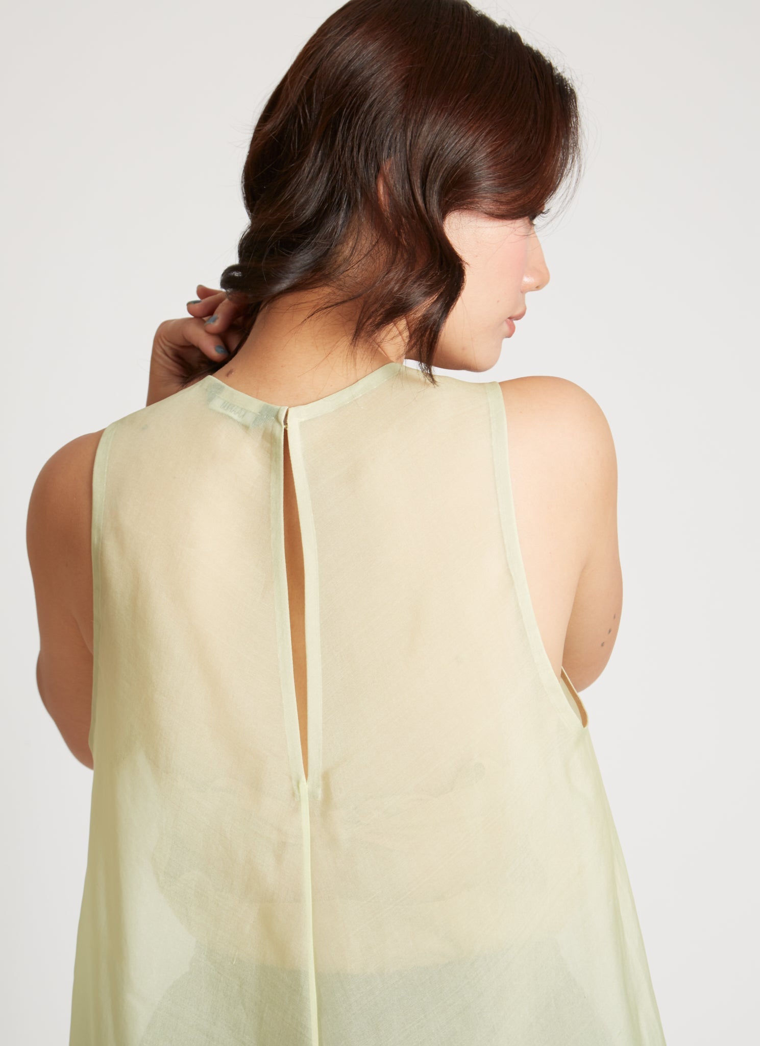 Close-up back view of a woman wearing chic and ethereal spring fashion in a Kaarem Green Rice Sleeveless Long Silk Organza Dress featuring a blind hem, button closure, and rear slit and is perfect for layering over slips or trousers. Each dress is crafted using traditional techniques to ensure lasting quality and movement at an Alas Eius store.