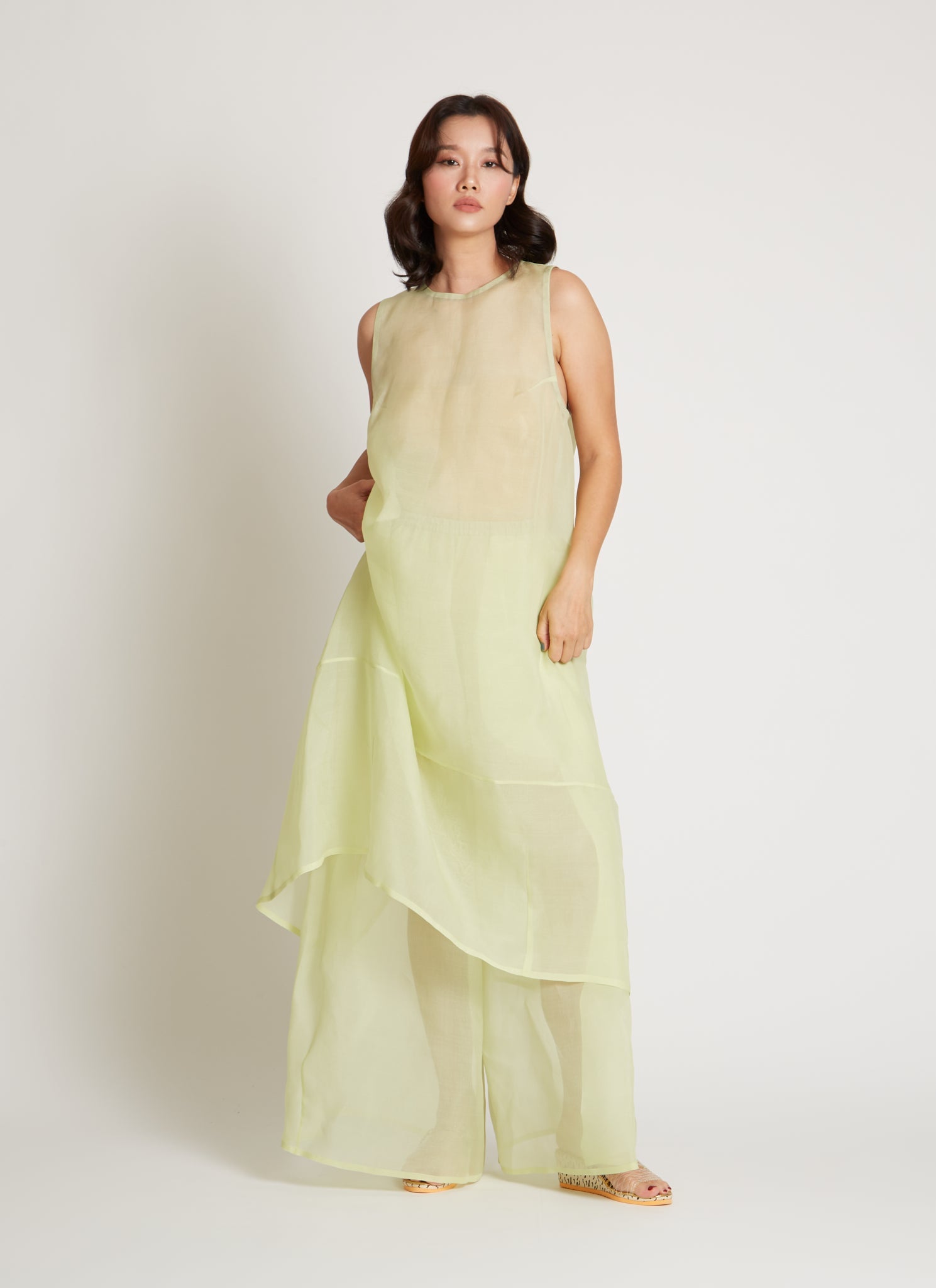 Full length front view of a woman wearing chic and ethereal spring fashion in a Kaarem Green Rice Sleeveless Long Silk Organza Dress featuring a blind hem, button closure, and rear slit and is perfect for layering over slips or trousers. Each dress is crafted using traditional techniques to ensure lasting quality and movement at an Alas Eius store.