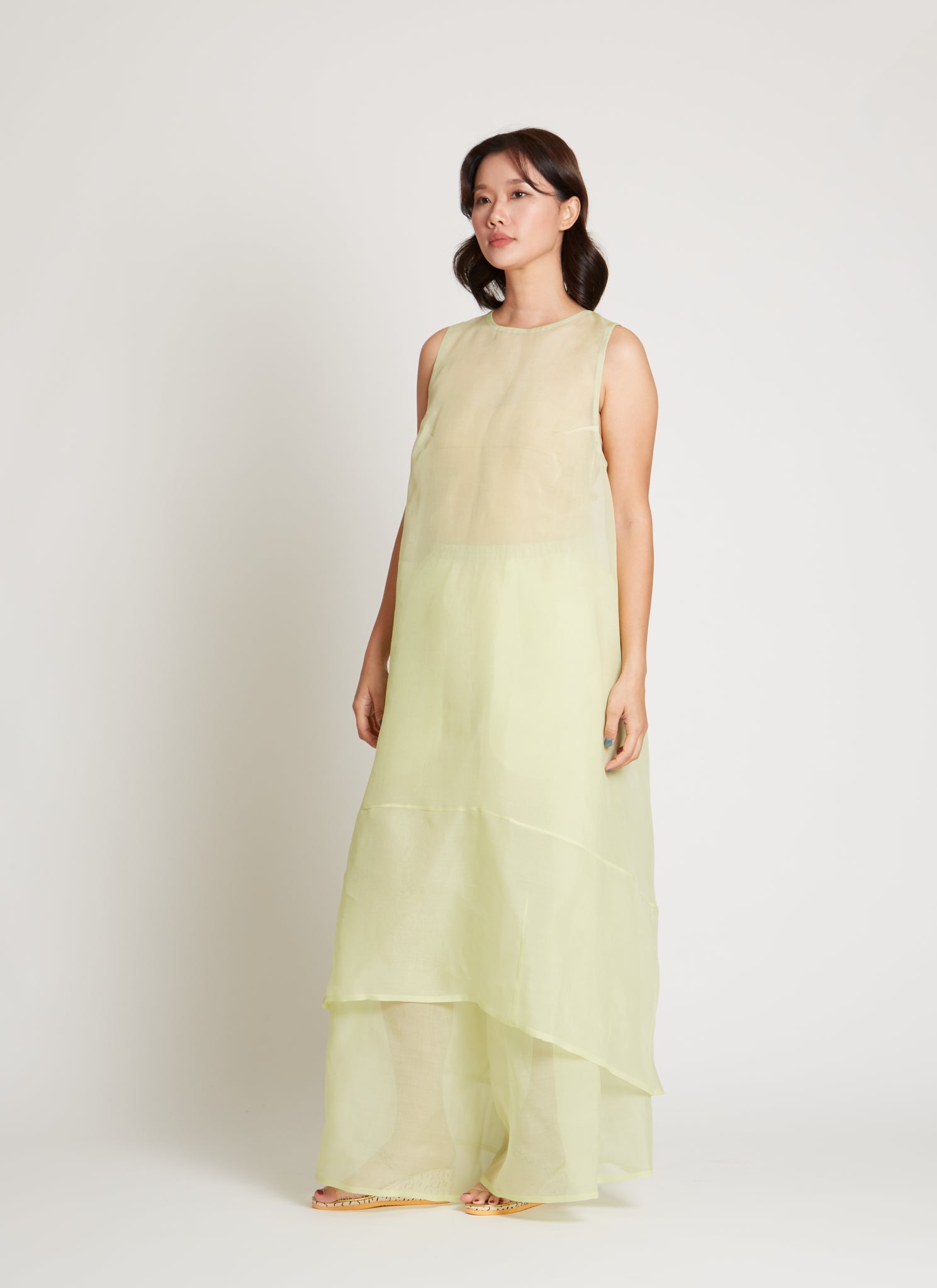 Full length front view of a woman wearing contemporary spring fashion in a Kaarem Green Rice Sleeveless Long Silk Organza Dress featuring a blind hem, button closure, and rear slit and is perfect for layering over slips or trousers. Each dress is crafted using traditional techniques to ensure lasting quality and movement at an Alas Eius store.