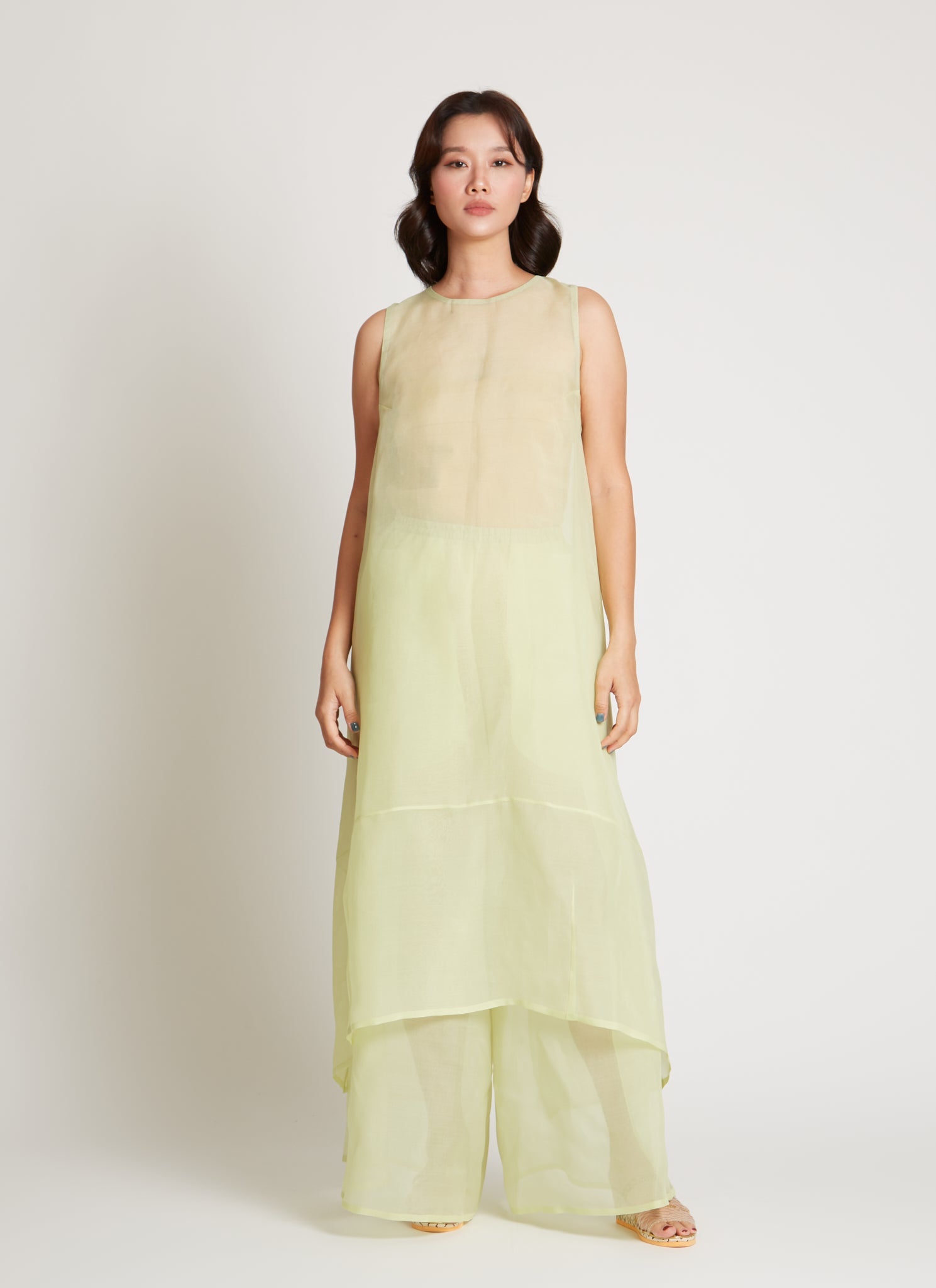 Full length front view of a woman wearing chic and ethereal spring fashion in a Kaarem Green Rice Sleeveless Long Silk Organza Dress featuring a blind hem, button closure, and rear slit and is perfect for layering over slips or trousers. Each dress is crafted using traditional techniques to ensure lasting quality and movement at an Alas Eius store.