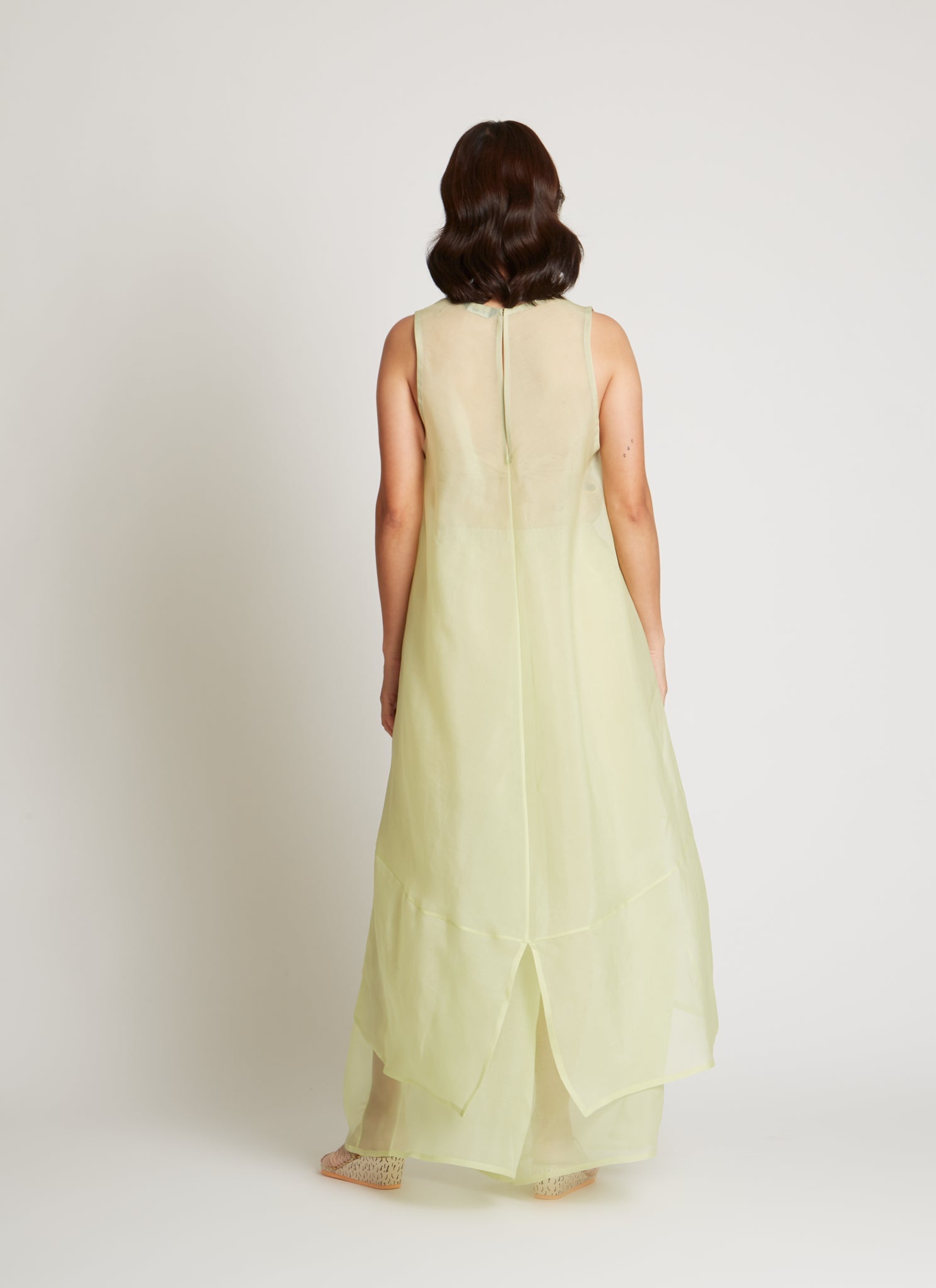 Full length back view of a woman wearing chic and ethereal spring fashion in a Kaarem Green Rice Sleeveless Long Silk Organza Dress featuring a blind hem, button closure, and rear slit and is perfect for layering over slips or trousers. Each dress is crafted using traditional techniques to ensure lasting quality and movement at an Alas Eius store.