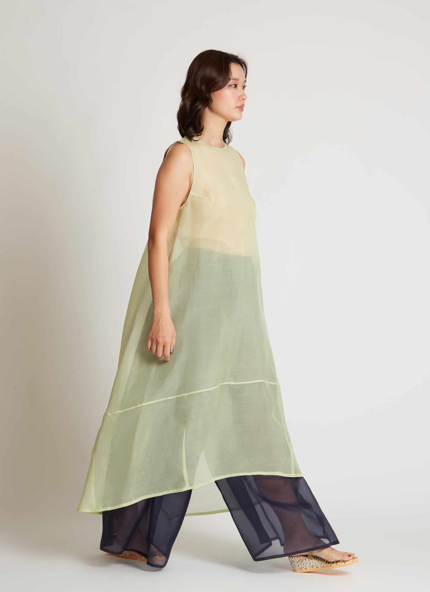 Woman in motion wearing chic and ethereal spring fashion in a Kaarem Green Rice Sleeveless Long Silk Organza Dress featuring a blind hem, button closure, and rear slit and is perfect for layering over slips or trousers. Each dress is crafted using traditional techniques to ensure lasting quality and movement at an Alas Eius store.