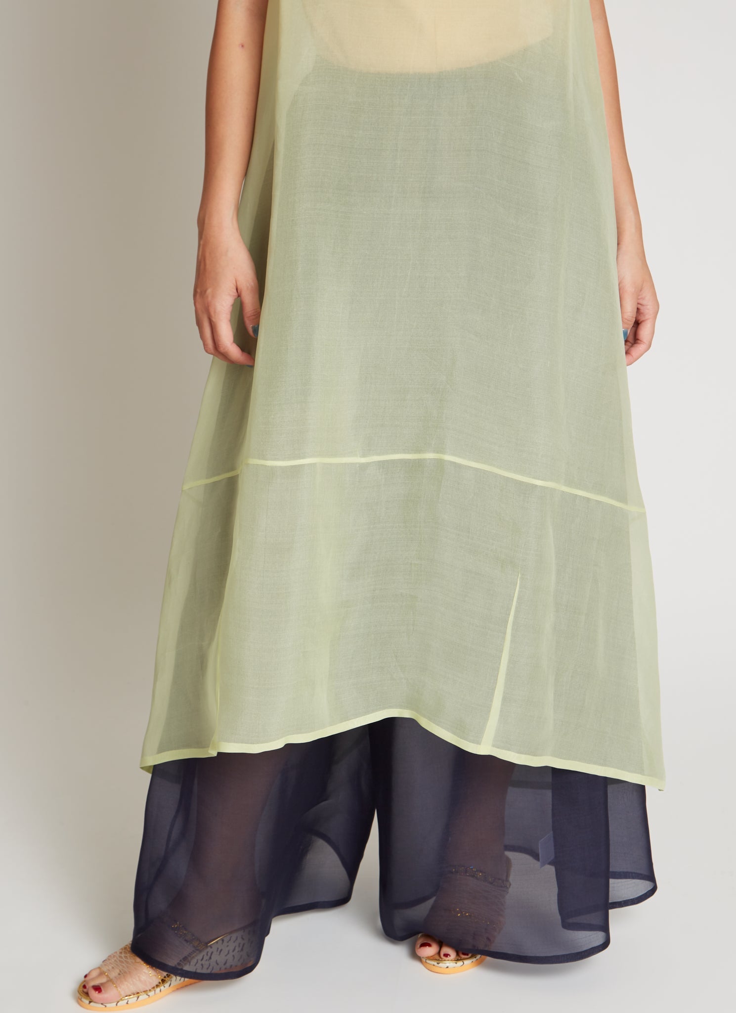 Close-up view of a woman wearing chic and ethereal spring fashion in a Kaarem Green Rice Sleeveless Long Silk Organza Dress featuring a blind hem, button closure, and rear slit and is perfect for layering over slips or trousers. Each dress is crafted using traditional techniques to ensure lasting quality and movement at an Alas Eius store.