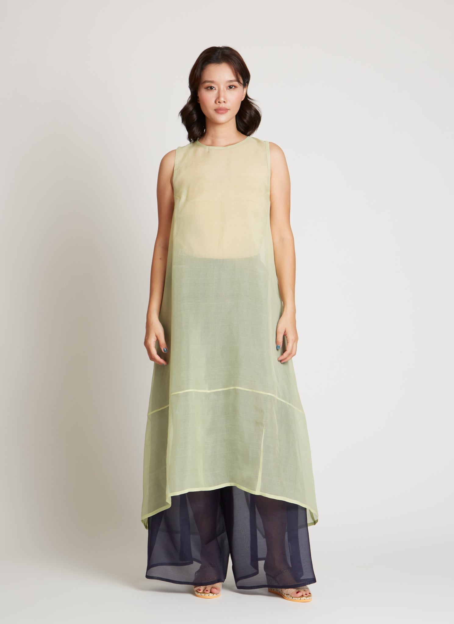 Full length front view of a woman wearing chic and ethereal spring fashion in a Kaarem Green Rice Sleeveless Long Silk Organza Dress featuring a blind hem, button closure, and rear slit and is perfect for layering over slips or trousers. Each dress is crafted using traditional techniques to ensure lasting quality and movement at an Alas Eius store.