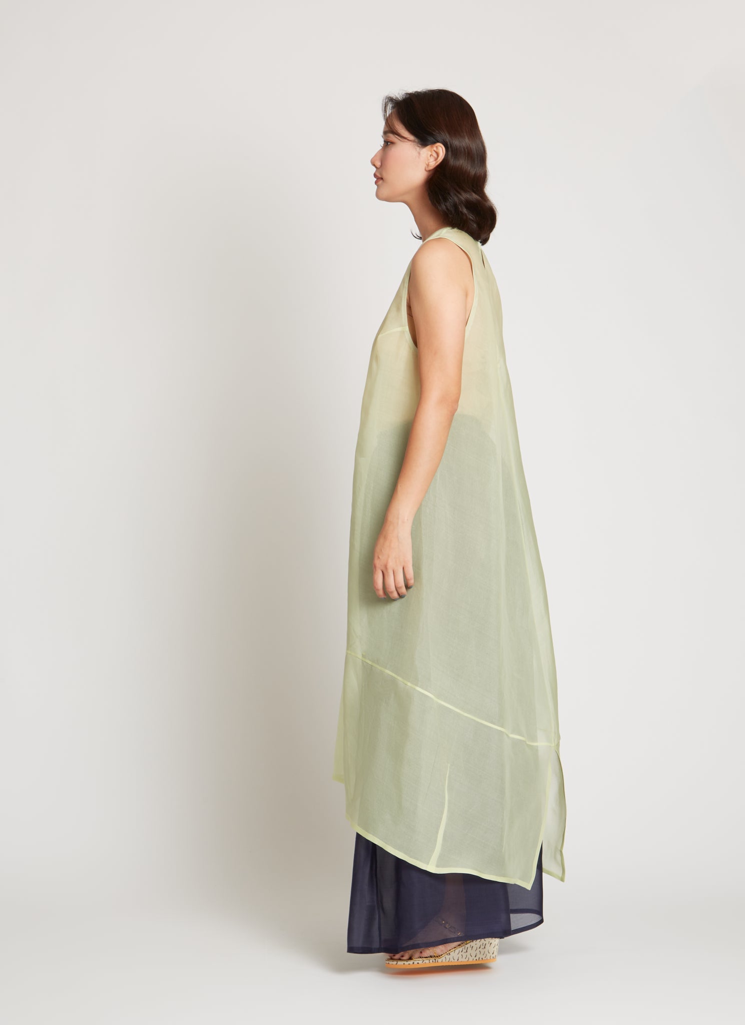 Full length side view of a woman wearing chic and ethereal spring fashion in a Kaarem Green Rice Sleeveless Long Silk Organza Dress featuring a blind hem, button closure, and rear slit and is perfect for layering over slips or trousers. Each dress is crafted using traditional techniques to ensure lasting quality and movement at an Alas Eius store.